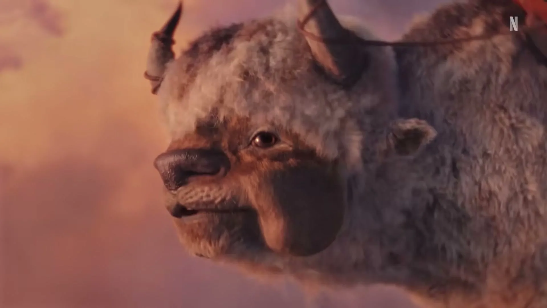 A large, furry, horned creature with a saddle on its back looks around with a neutral expression against a sunset sky.