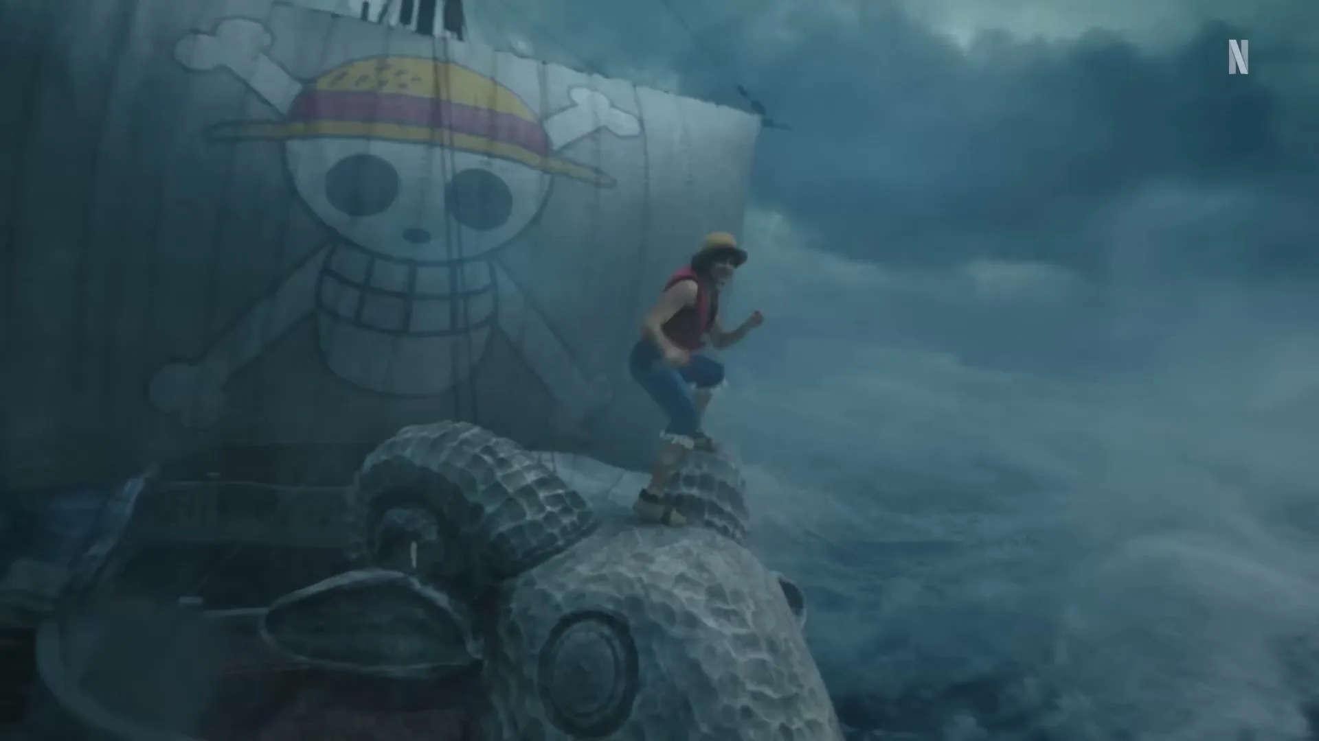 A young man in a straw hat stands on the bow of a ship with a skull and crossbones sail, battling rough seas under a stormy sky.