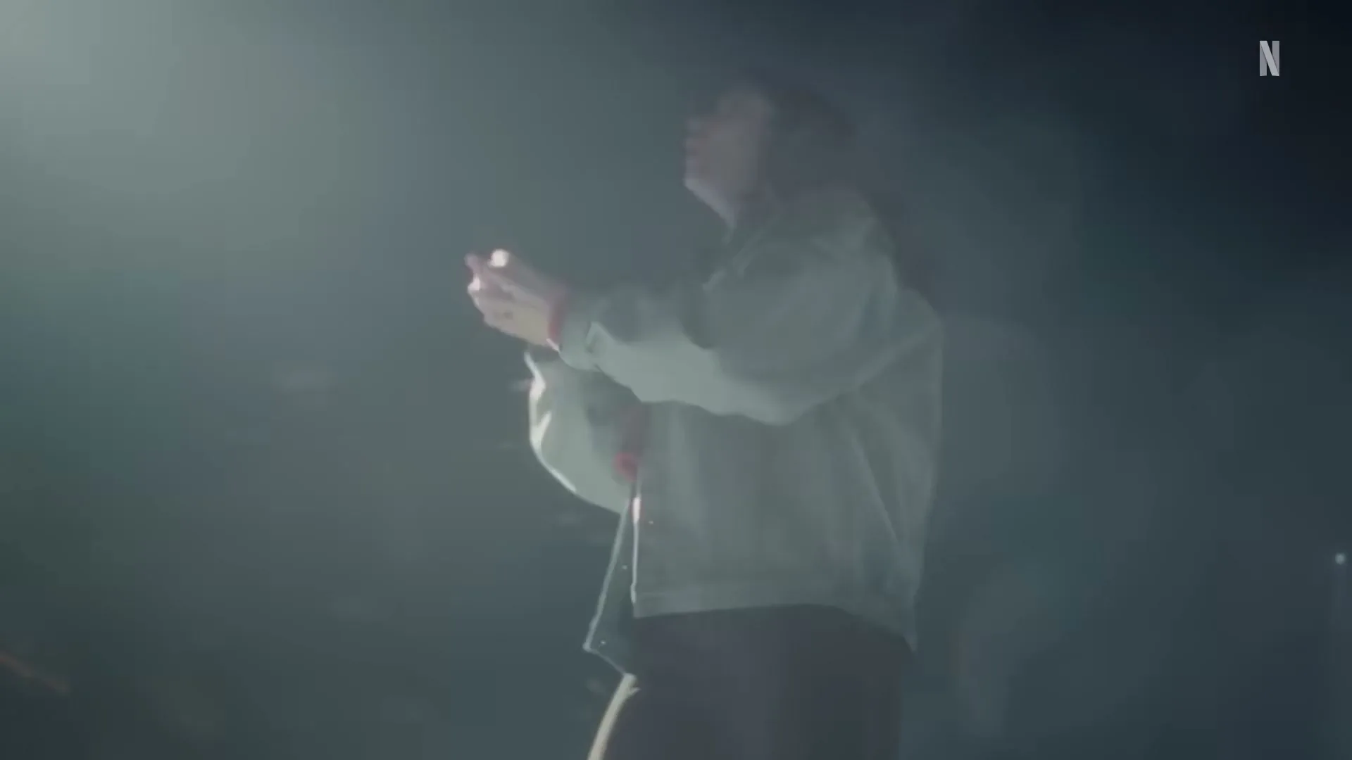 A young woman with long hair, wearing a light jacket, stands in a smoky, dark space, looking up and moving her hands.