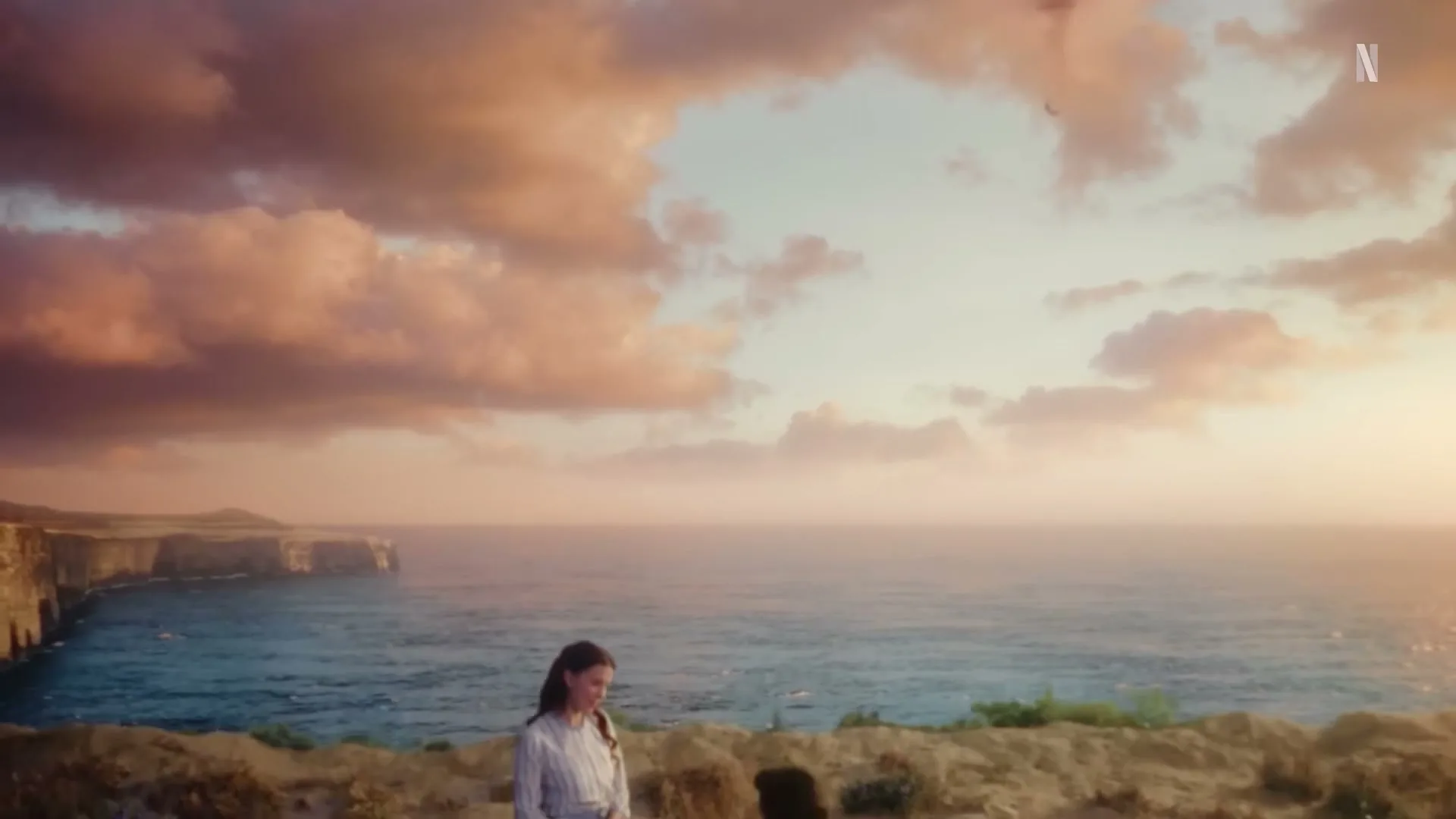 A young woman in a period dress stands on a cliff overlooking the ocean as a man kneels before her, proposing marriage at sunset.