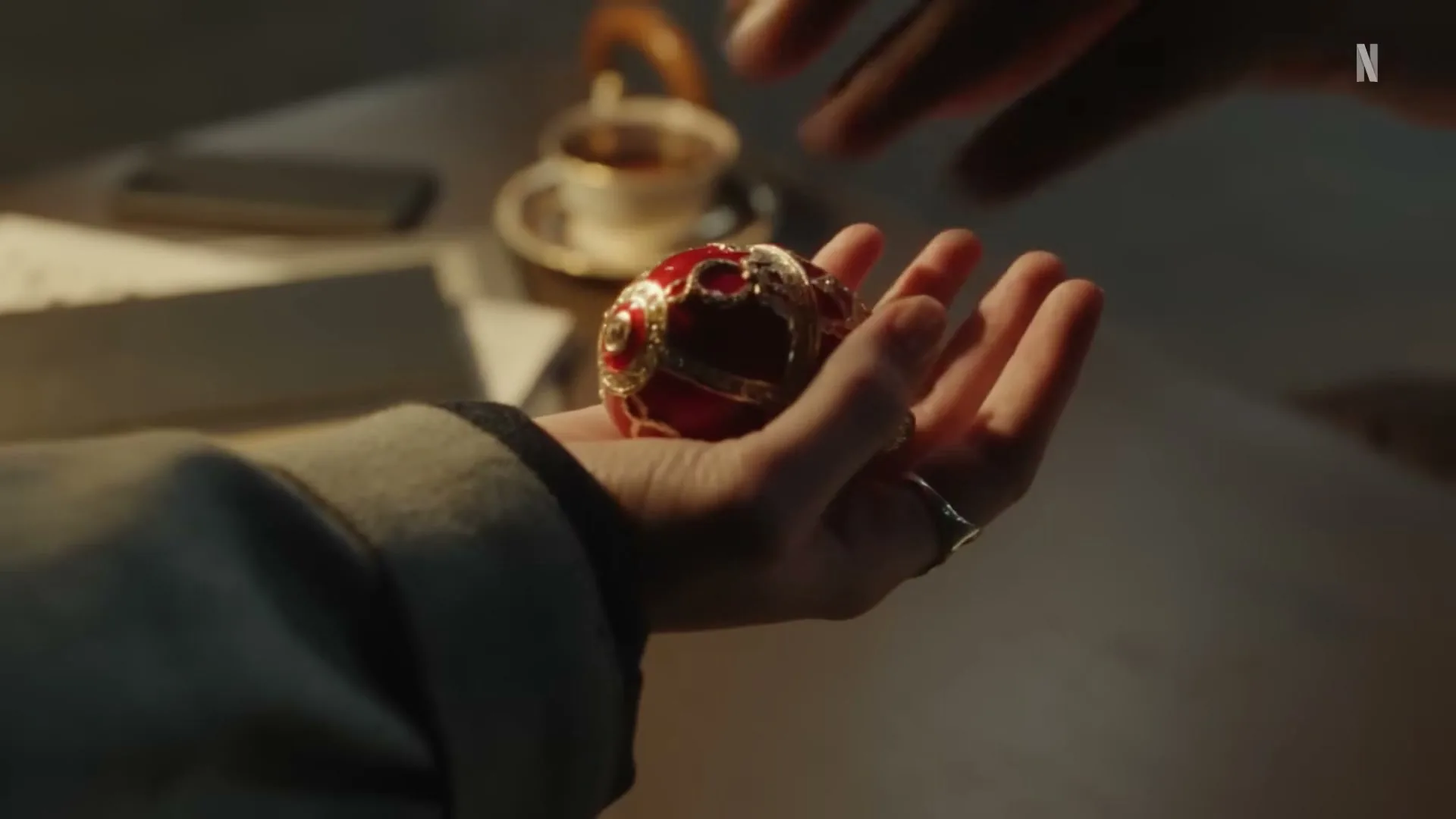 A person's hand holds a red ornate egg, then another hand reaches in to take it, on a table with a teacup.