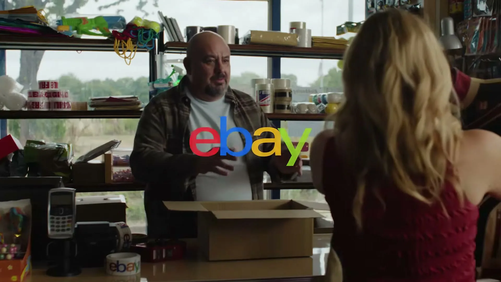 A bald man in a plaid shirt stands behind a counter, talking to a blonde woman in a red top, as they pack an item into a box in a cluttered store.