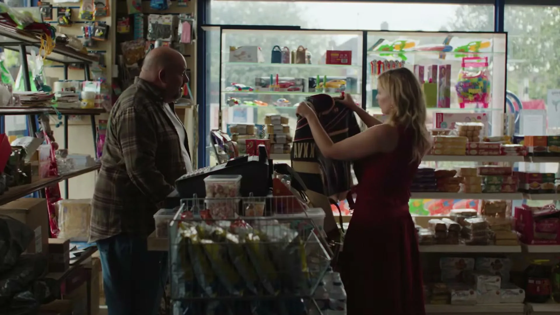 A woman in a red dress shows a jacket to a male shopkeeper in a small, cluttered convenience store.