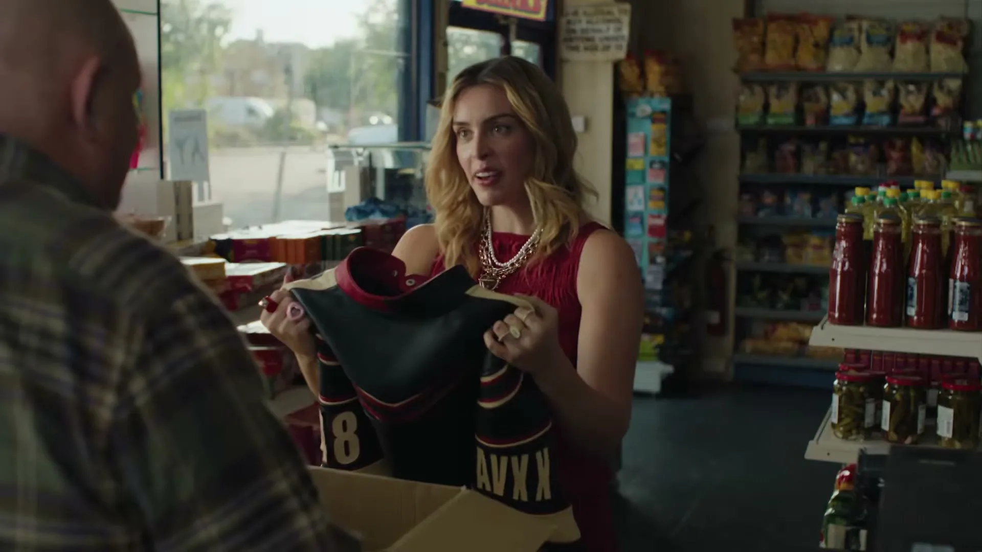 A woman in a red dress and gold jewelry holds up a black and red jersey with the number 8 and "AVXX" on it, showing it to a man in a plaid shirt in a convenience store.