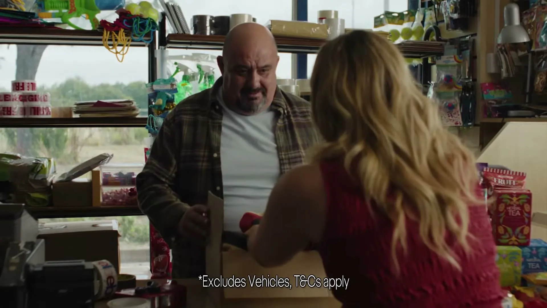 A bald man in a plaid shirt watches a blonde woman in a red top pack items into a box at a counter in a shop.