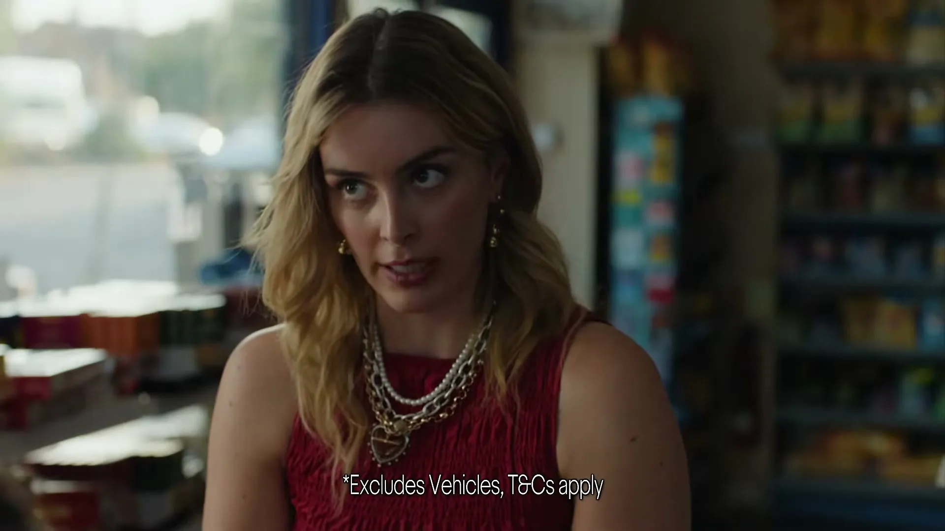 A young woman with blonde hair and a red top looks directly at the camera with a slightly annoyed expression in a convenience store.