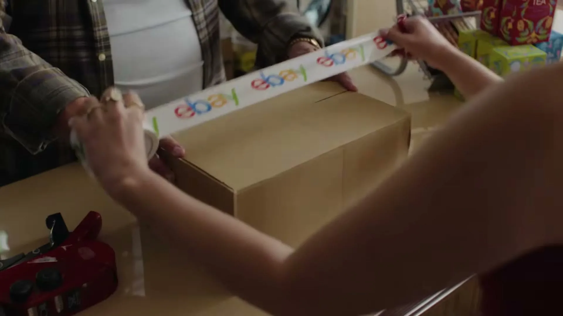 Two people's hands are shown taping up a cardboard box with eBay branded tape on a counter.