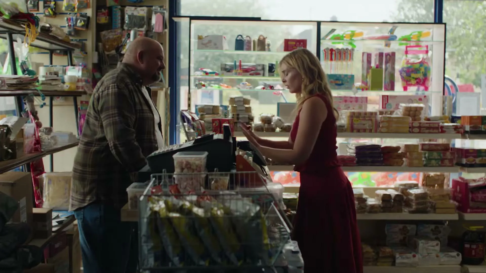 A blonde woman in a red dress stands at a counter in a convenience store, looking at a male cashier.