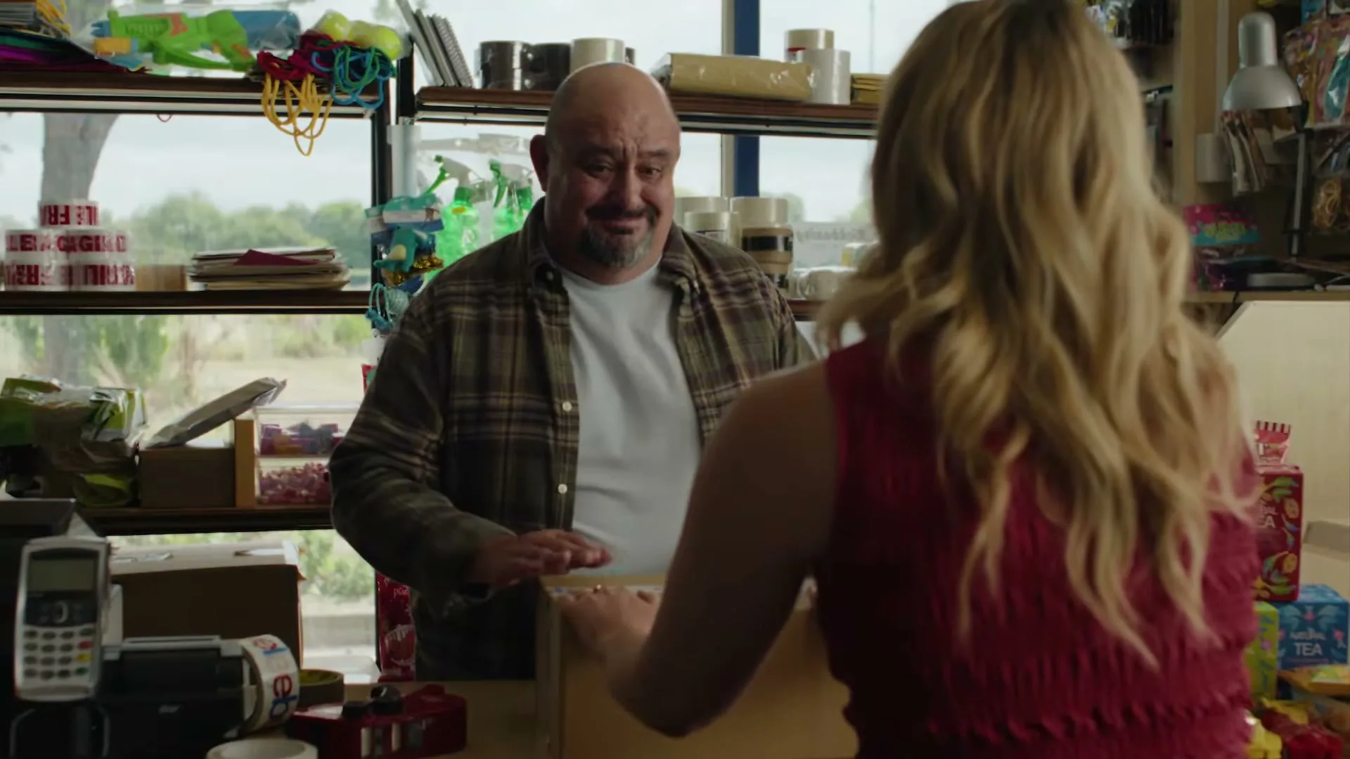 A bald man in a plaid shirt smiles at a blonde woman in a red top as she packs a box on a counter in a small shop.