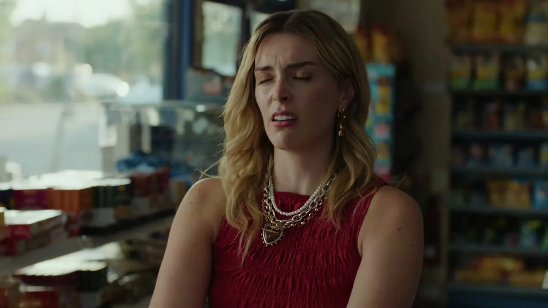 A blonde woman in a red dress and layered necklaces stands in a convenience store, looking pained with her eyes closed.
