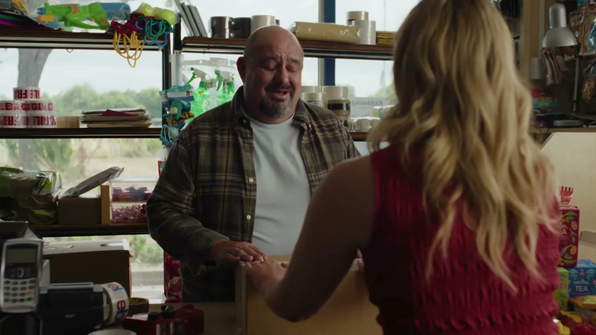 A bald man in a plaid shirt smiles at a blonde woman in a red top, who is packaging a box on a counter in a shop.