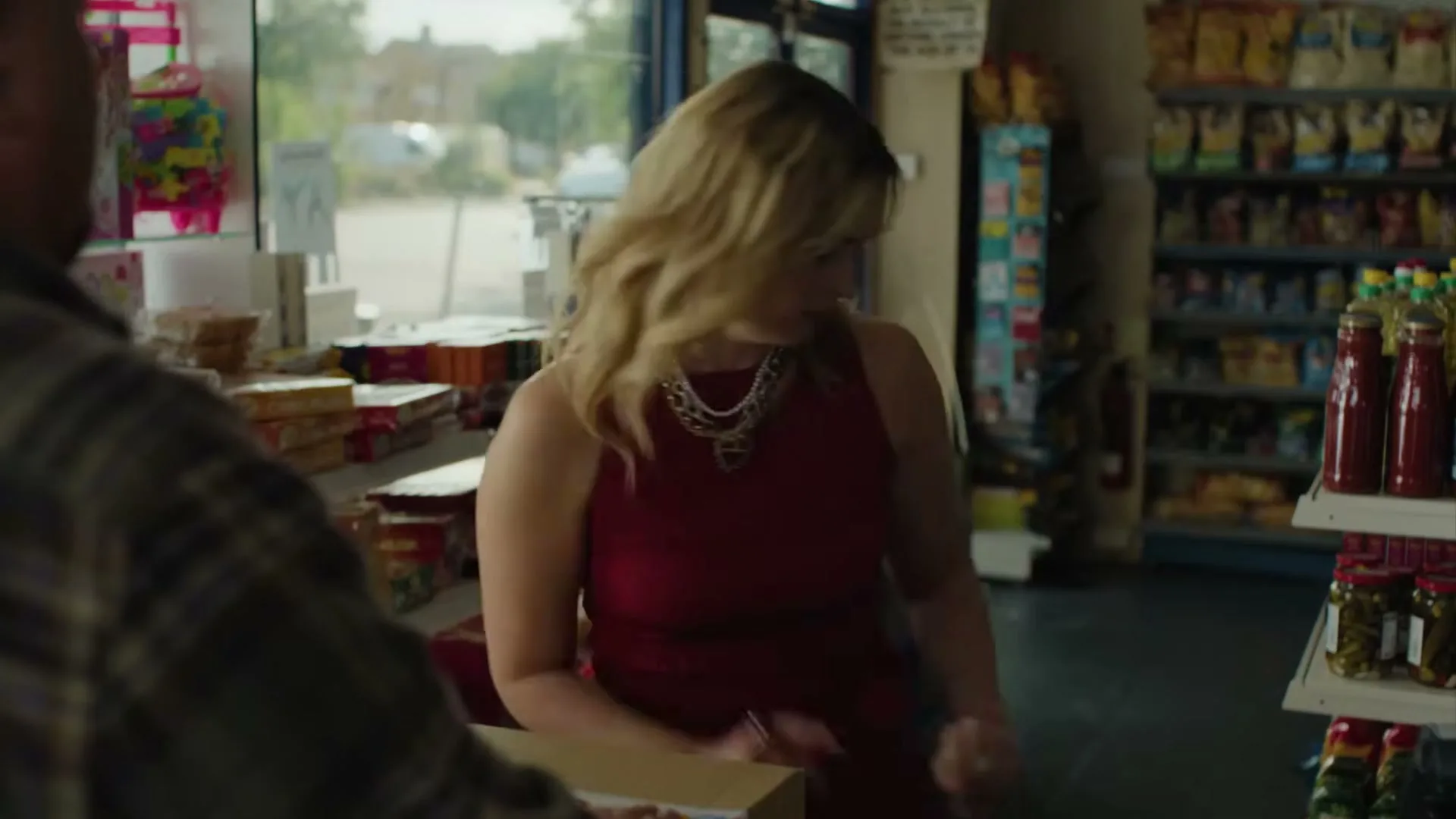 A woman in a red dress and pearl necklace looks at a man across a counter in a convenience store, then bends down out of frame.