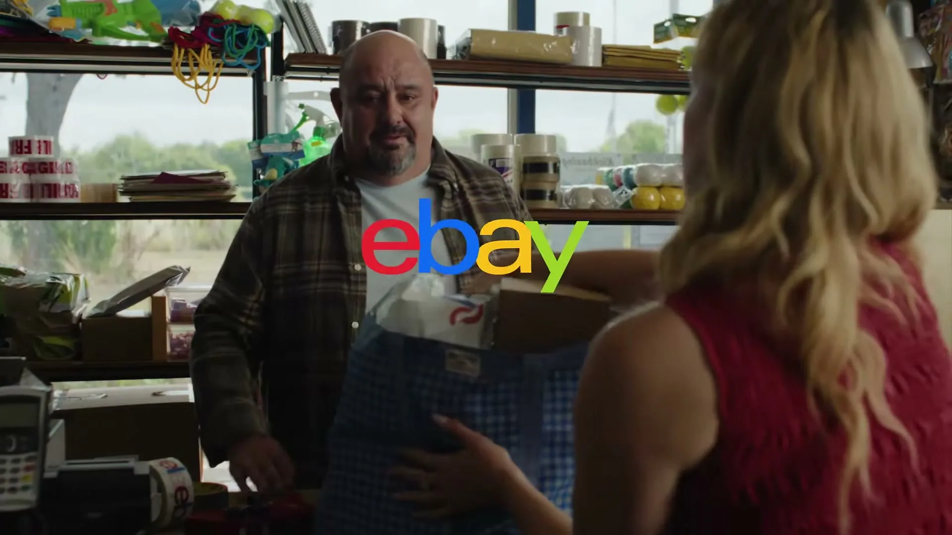 A bald man in a plaid shirt smiles at a blonde woman in a red top who is holding up a blue checkered shirt in a cluttered shop.
