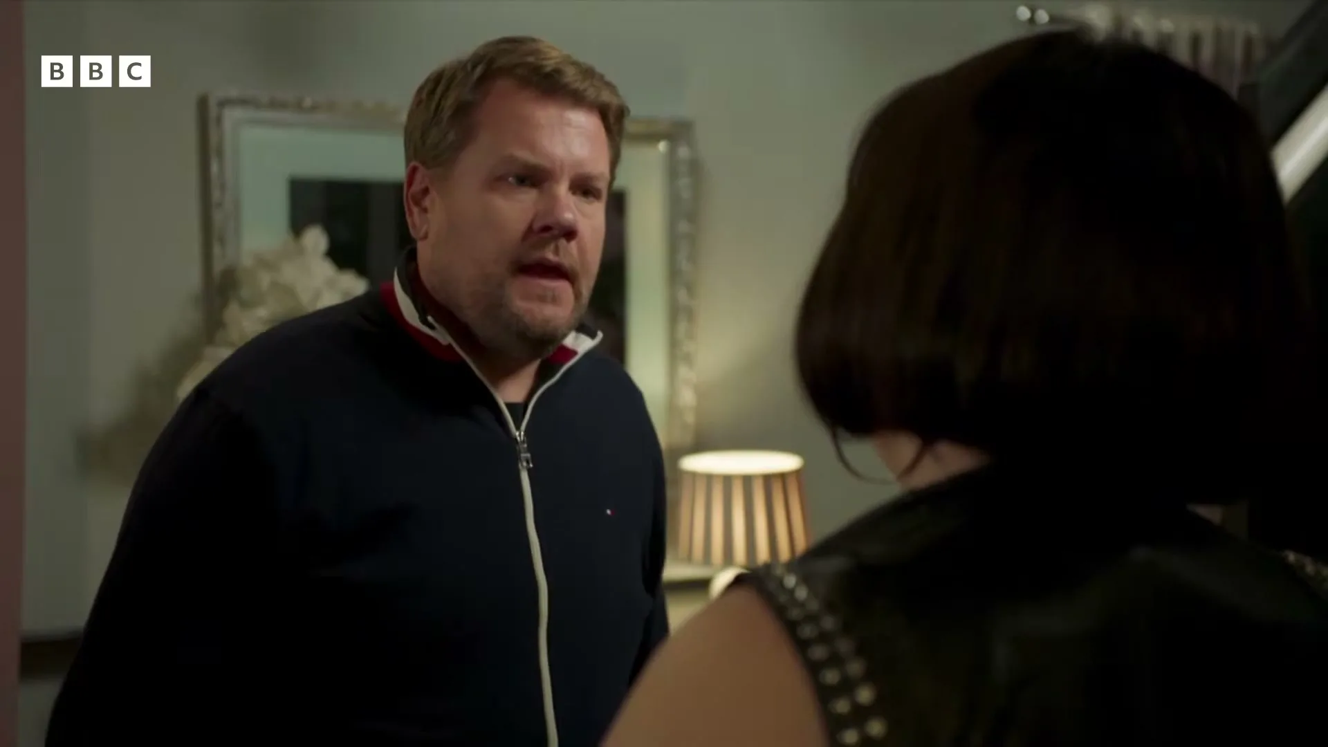 James Corden, a male in his 40s, wearing a navy blue jacket, looks concerned while talking to a woman with her back to the camera in a dimly lit interior.