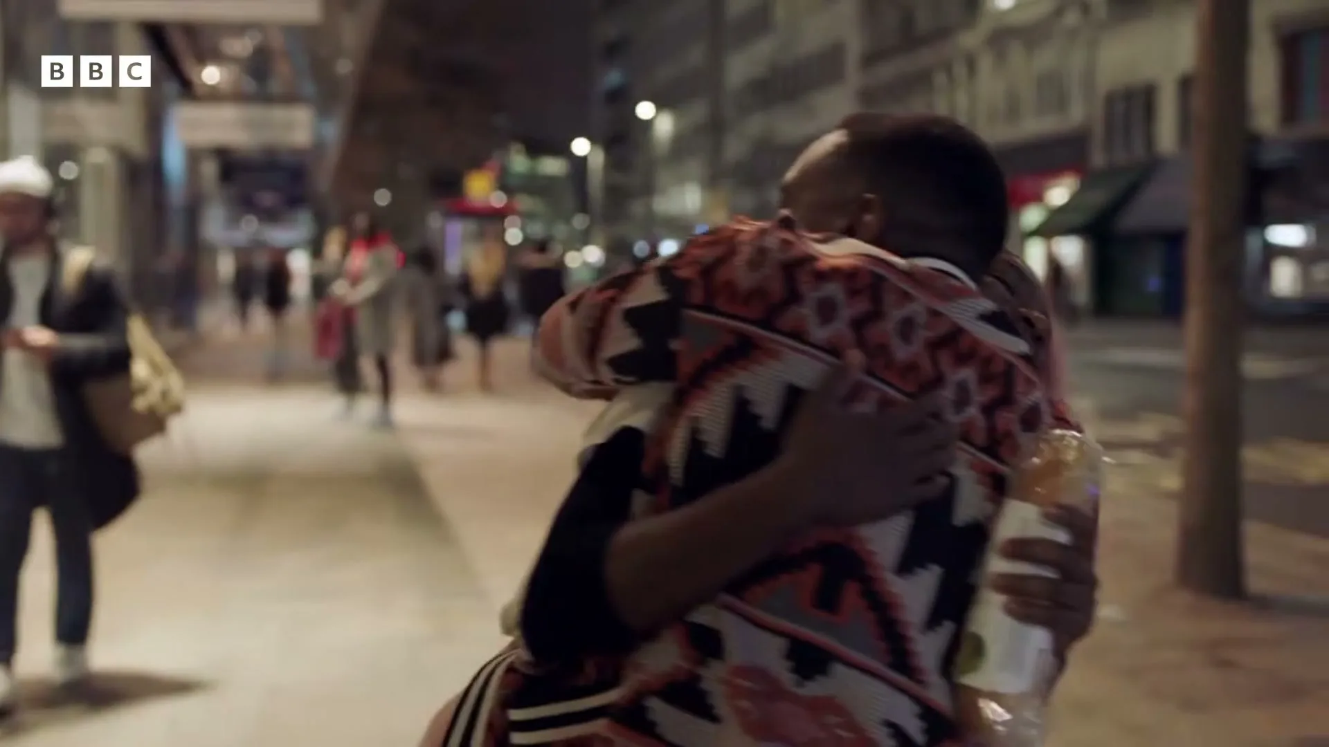 Two young adults, a man and a woman with pink hair, embrace on a busy city sidewalk at night, holding a wrapped sandwich.