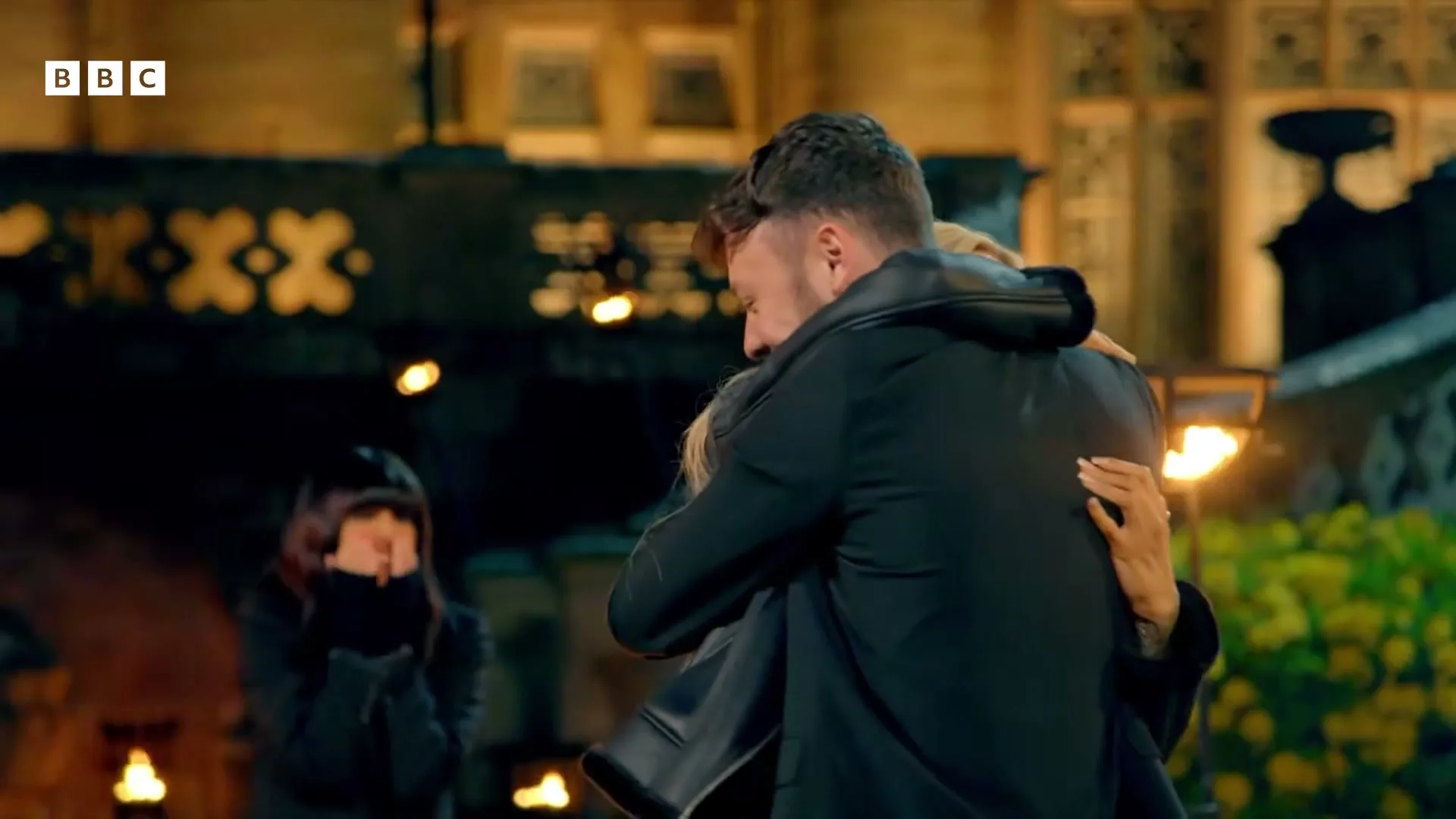 A man and a woman embrace tightly, while another woman in the background watches with her hands covering her mouth, at night outside a building.