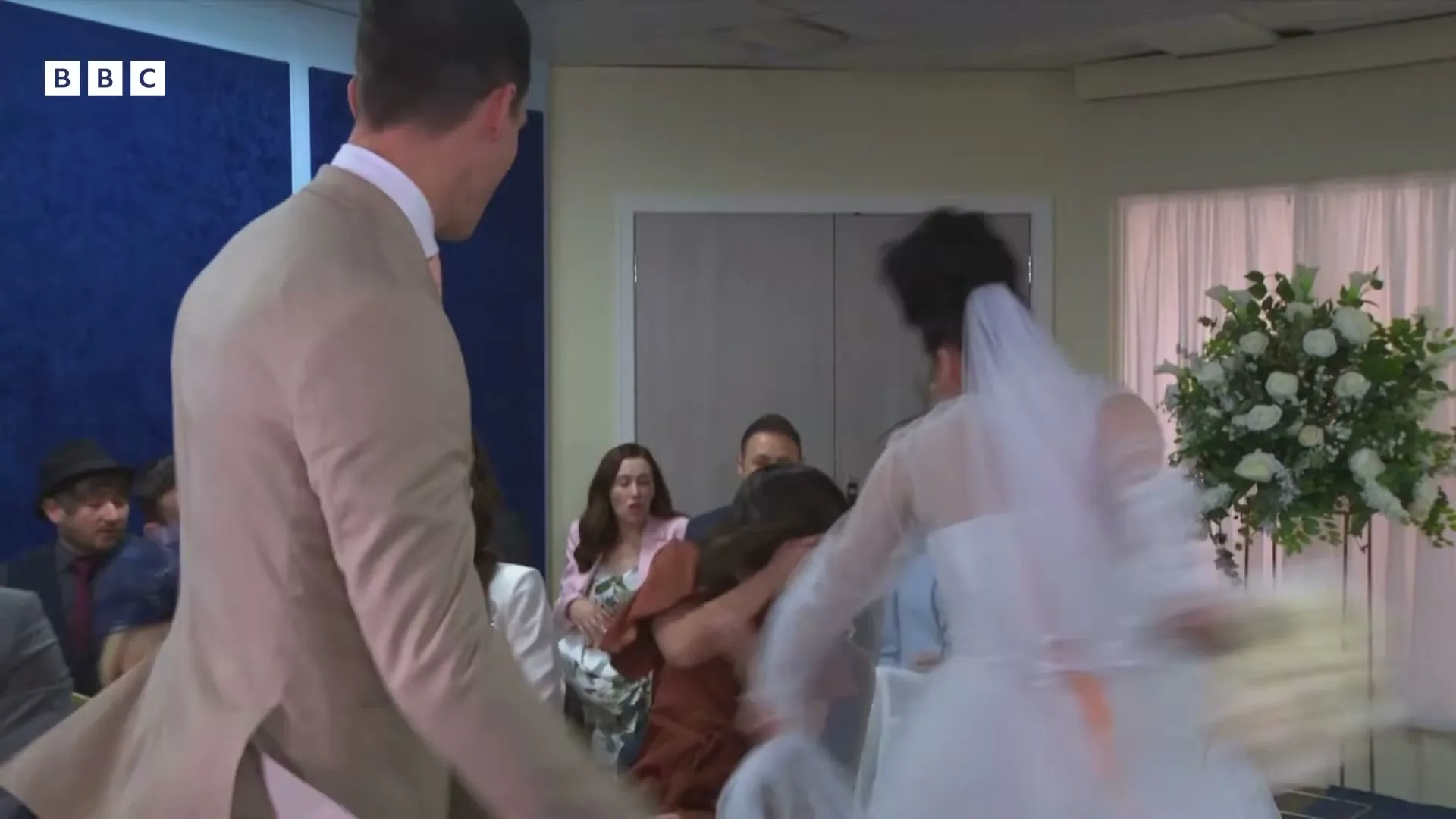 A groom in a beige suit and a bride in a white dress are seen from behind, walking down the aisle at a wedding ceremony, with guests seated on either side.