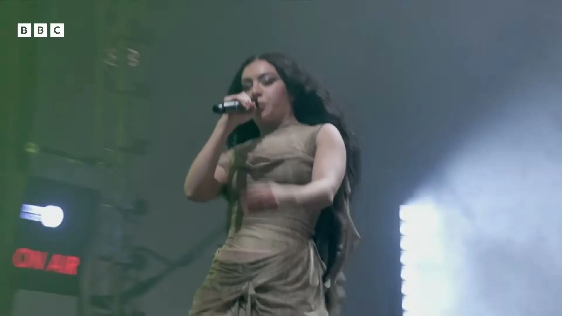 A female singer with long dark hair performs on stage, holding a microphone and wearing a sheer, draped dress.