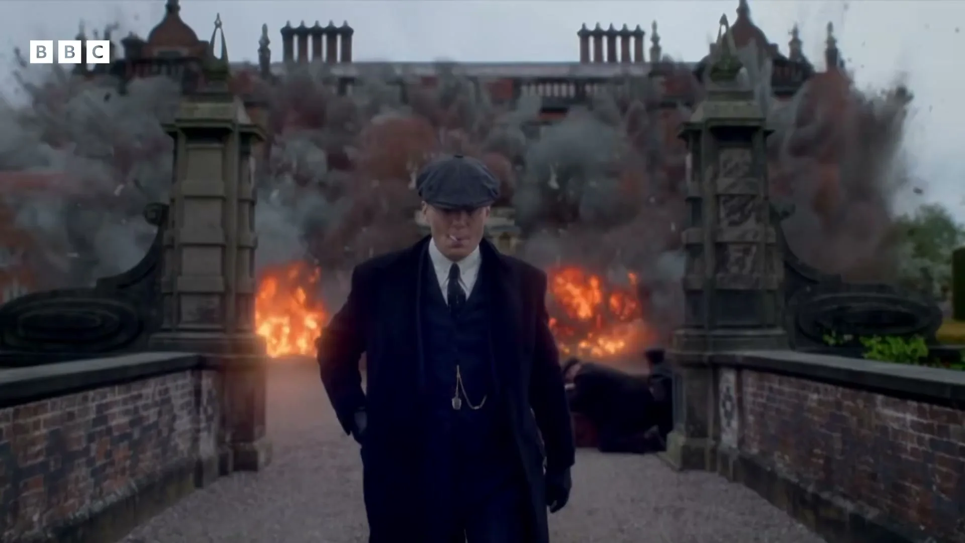 A man in a suit and flat cap walks away from a large manor house engulfed in explosions and fire, looking calm and smoking a cigarette.