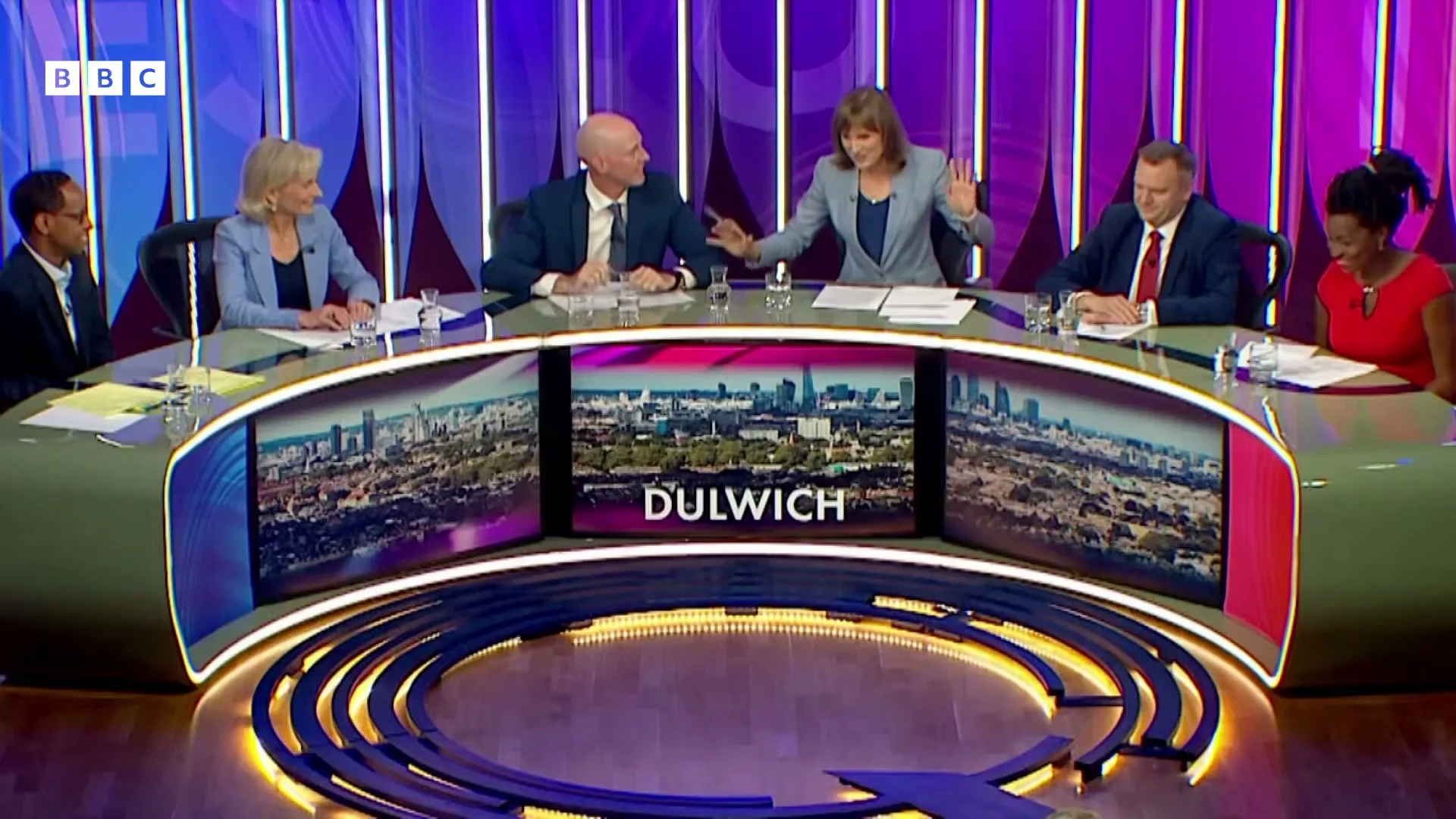Six panelists, three men and three women, sit around a curved desk with screens displaying "DULWICH" and cityscapes, in a brightly lit studio.