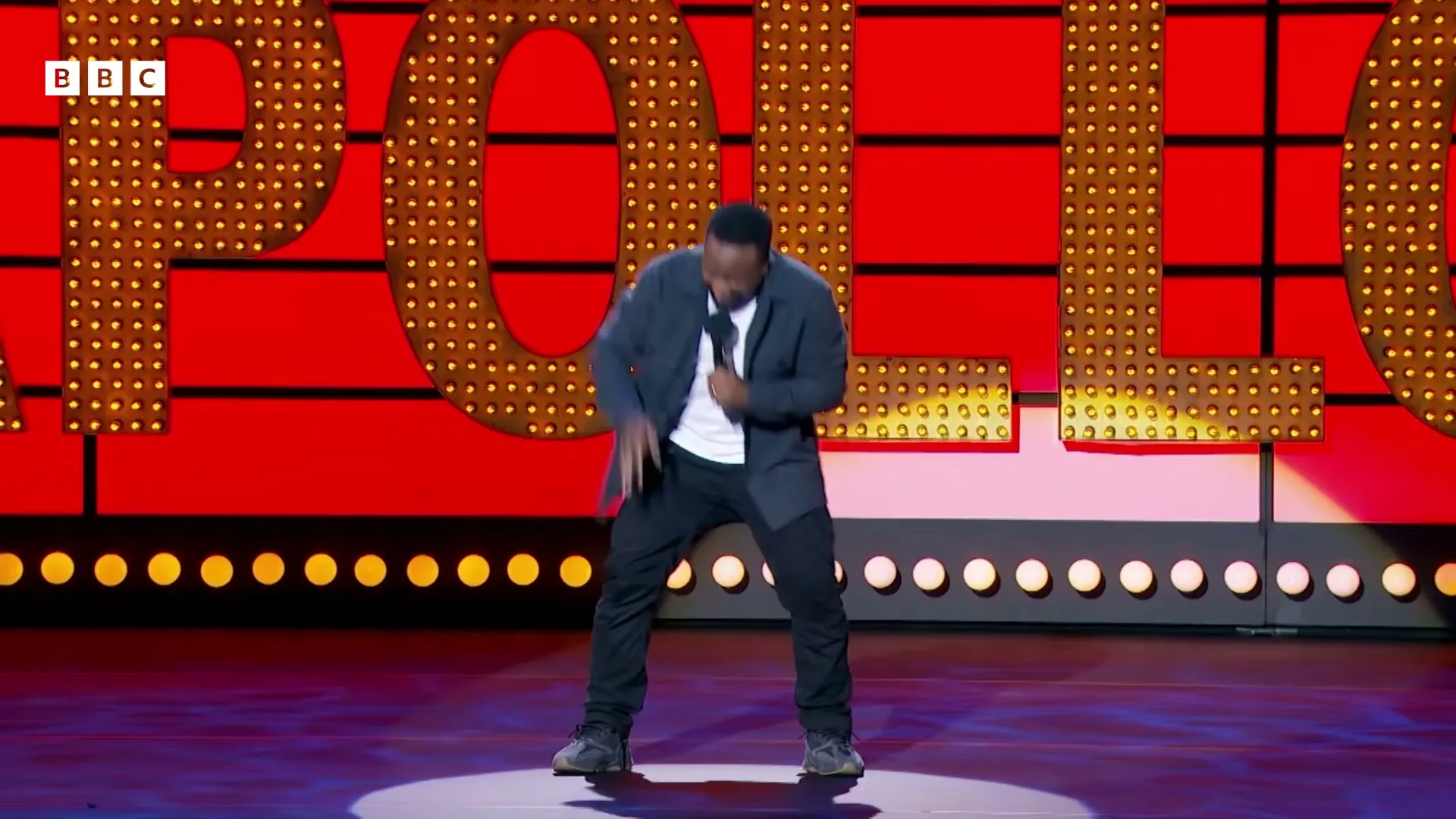 A Black male comedian in a dark jacket and white t-shirt performs on a brightly lit stage with a large "APOLLO" sign in the background.