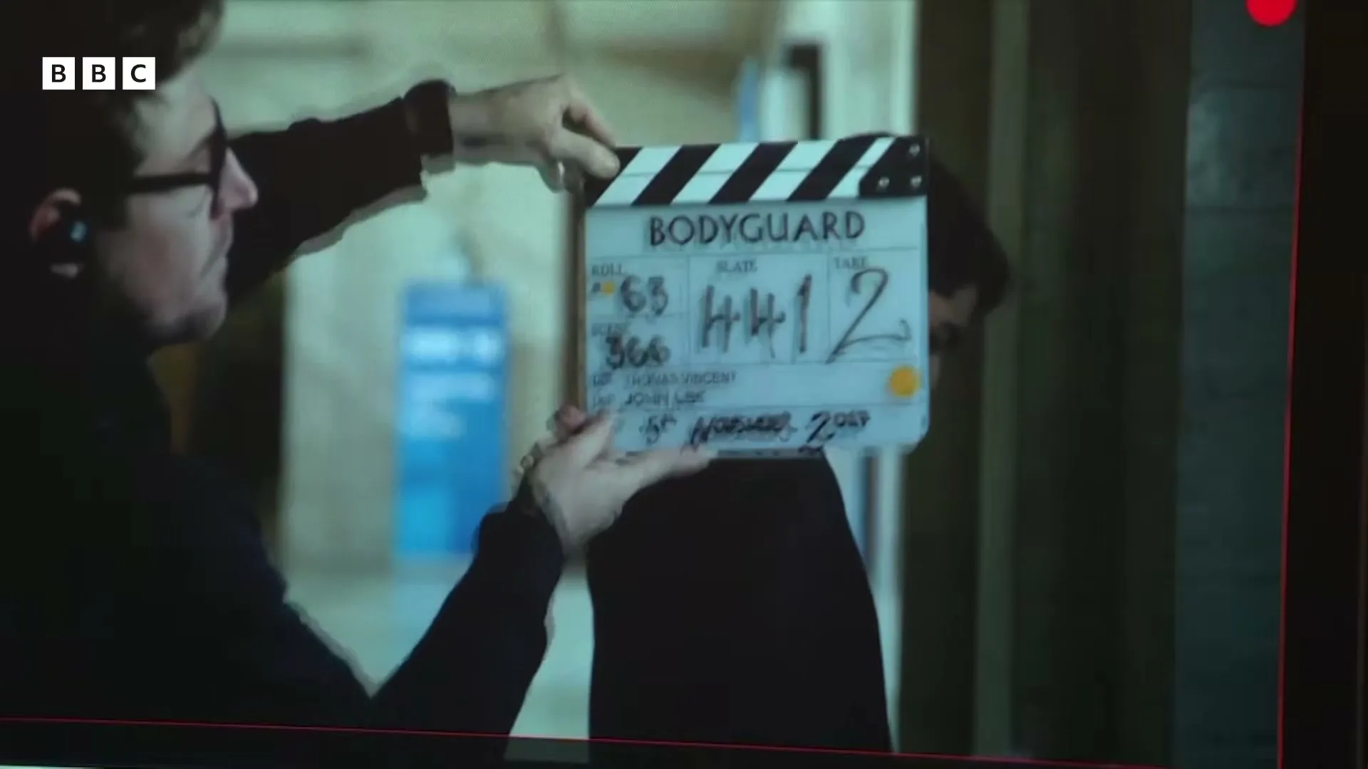 A man in glasses holds a clapperboard for "Bodyguard" on a film set, with another person blurred in the background.