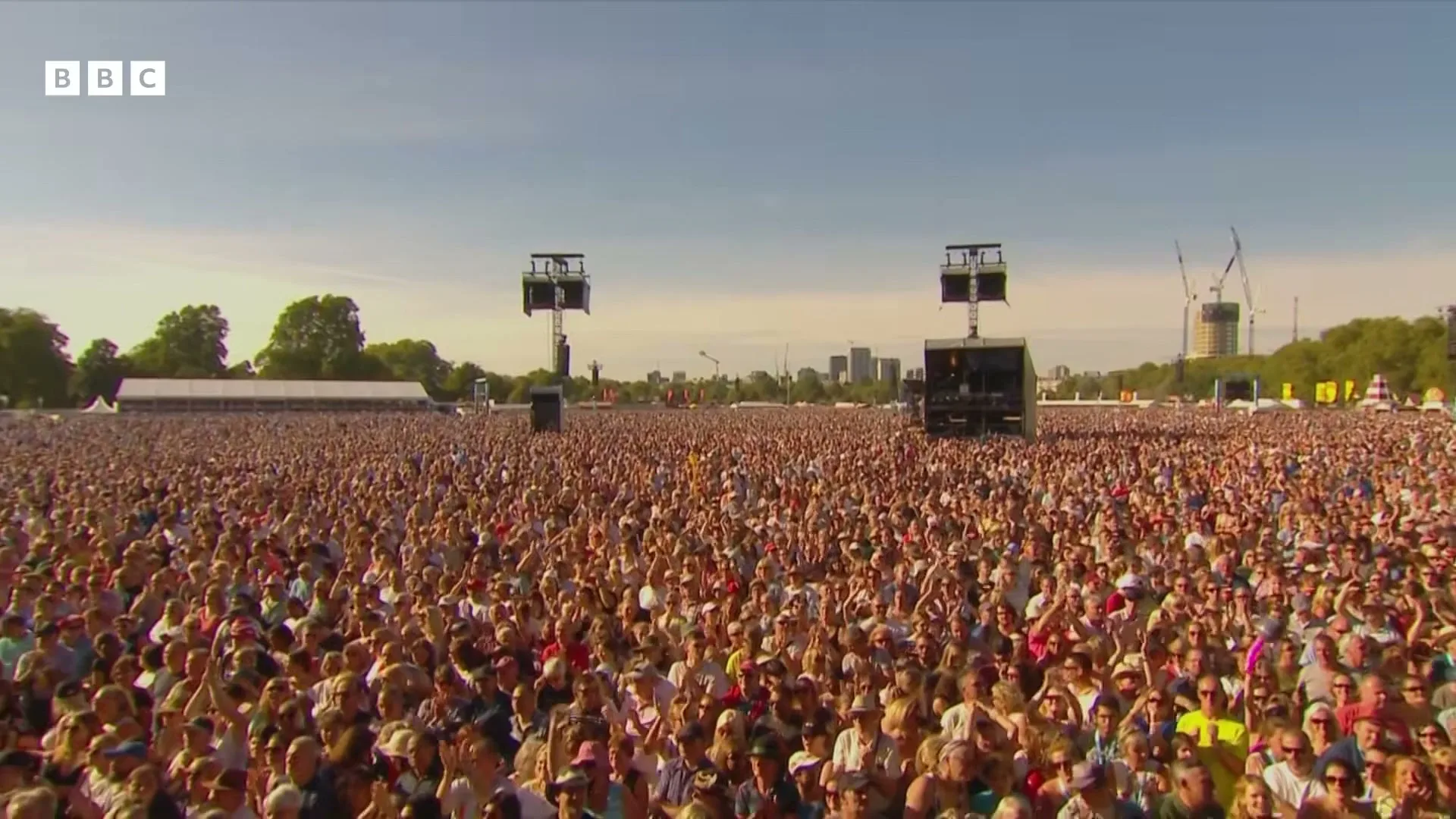 A massive crowd of people, mostly adults, are seen from a high angle, cheering and clapping at an outdoor music festival on a sunny day.