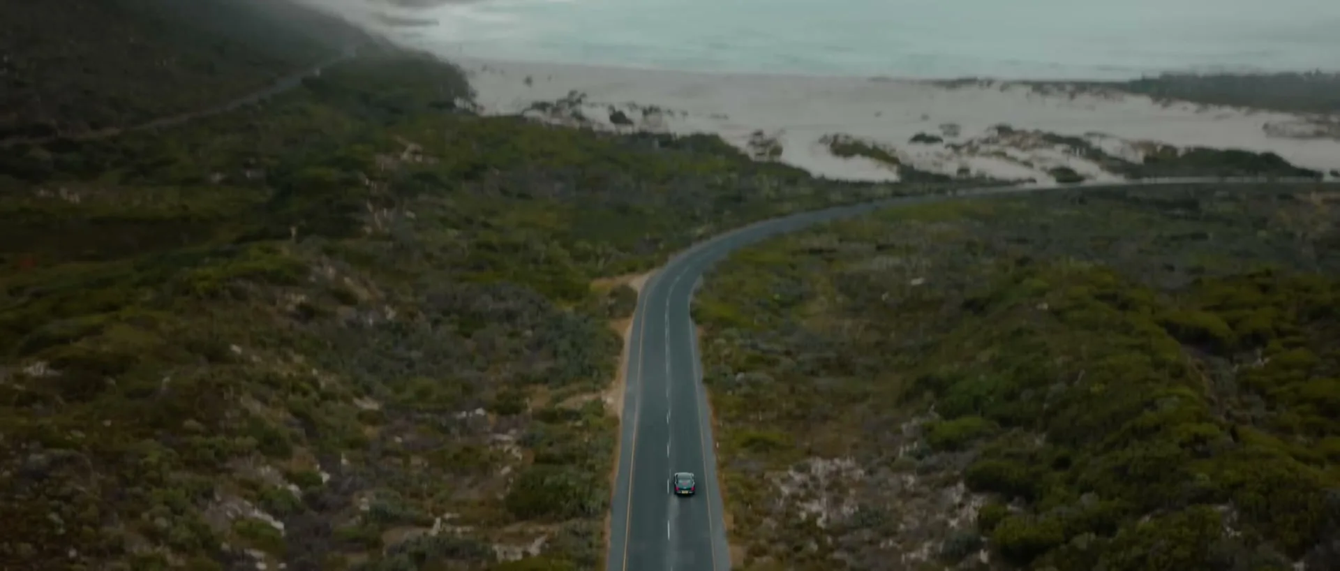 An aerial shot of a dark blue Bentley driving on a winding coastal road, surrounded by green bushes and a sandy beach under a cloudy sky.