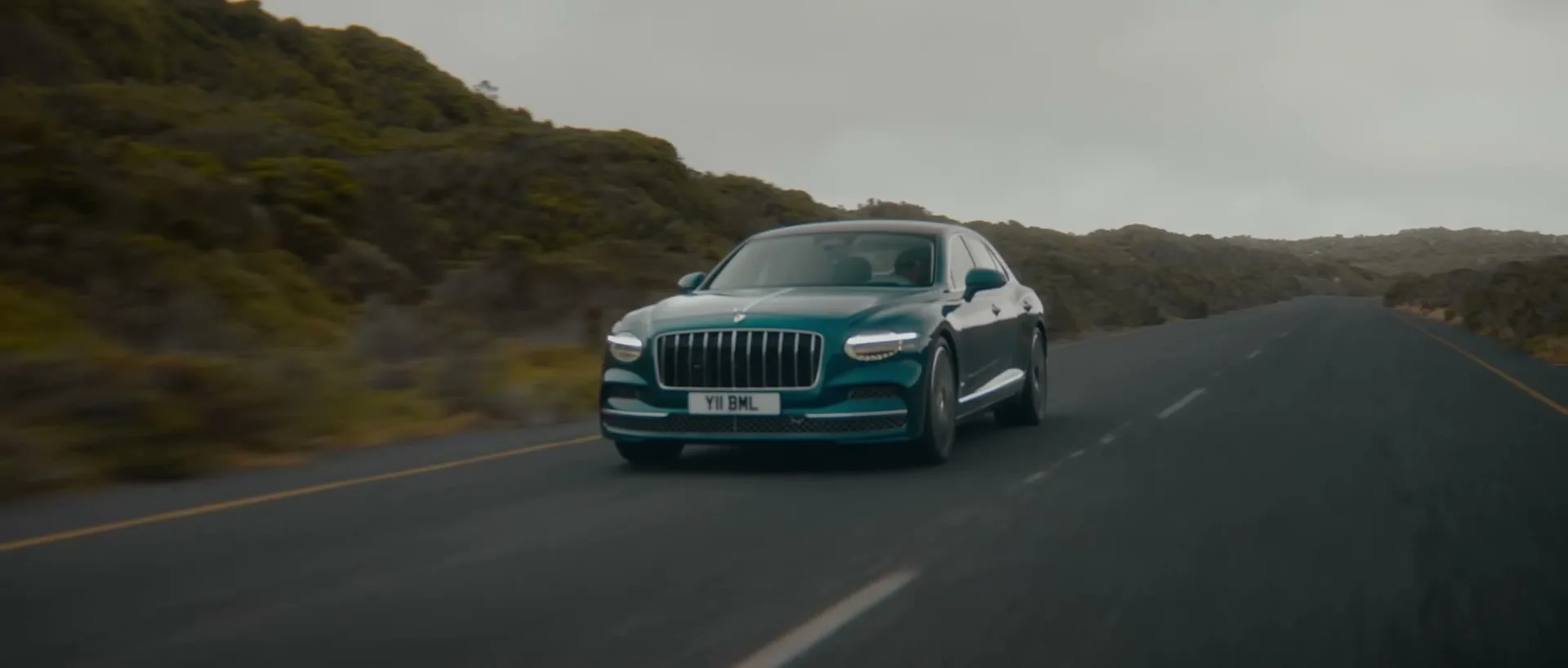 A teal Bentley car drives on a winding road through a mountainous, overcast landscape.