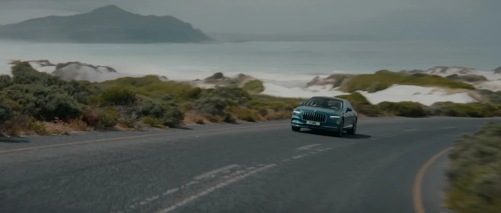 A teal Bentley car drives on a coastal road with sand dunes and the ocean in the background under an overcast sky.