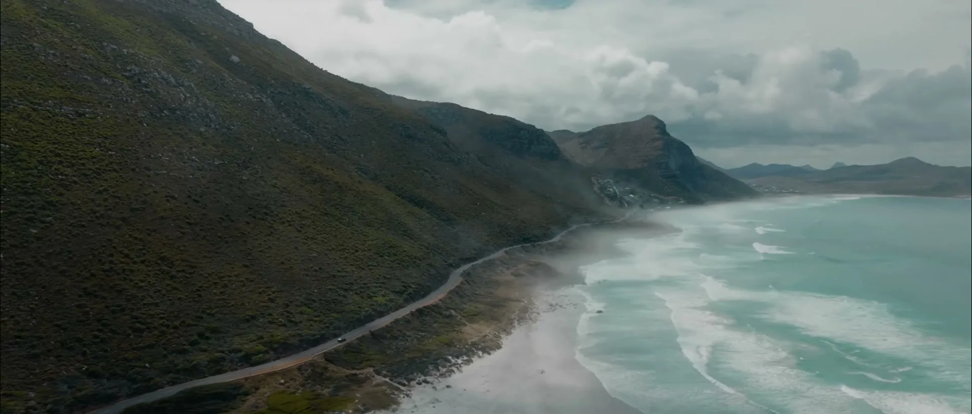 An aerial view shows a car driving on a coastal road next to a beach with waves crashing, surrounded by mountains under a cloudy sky.