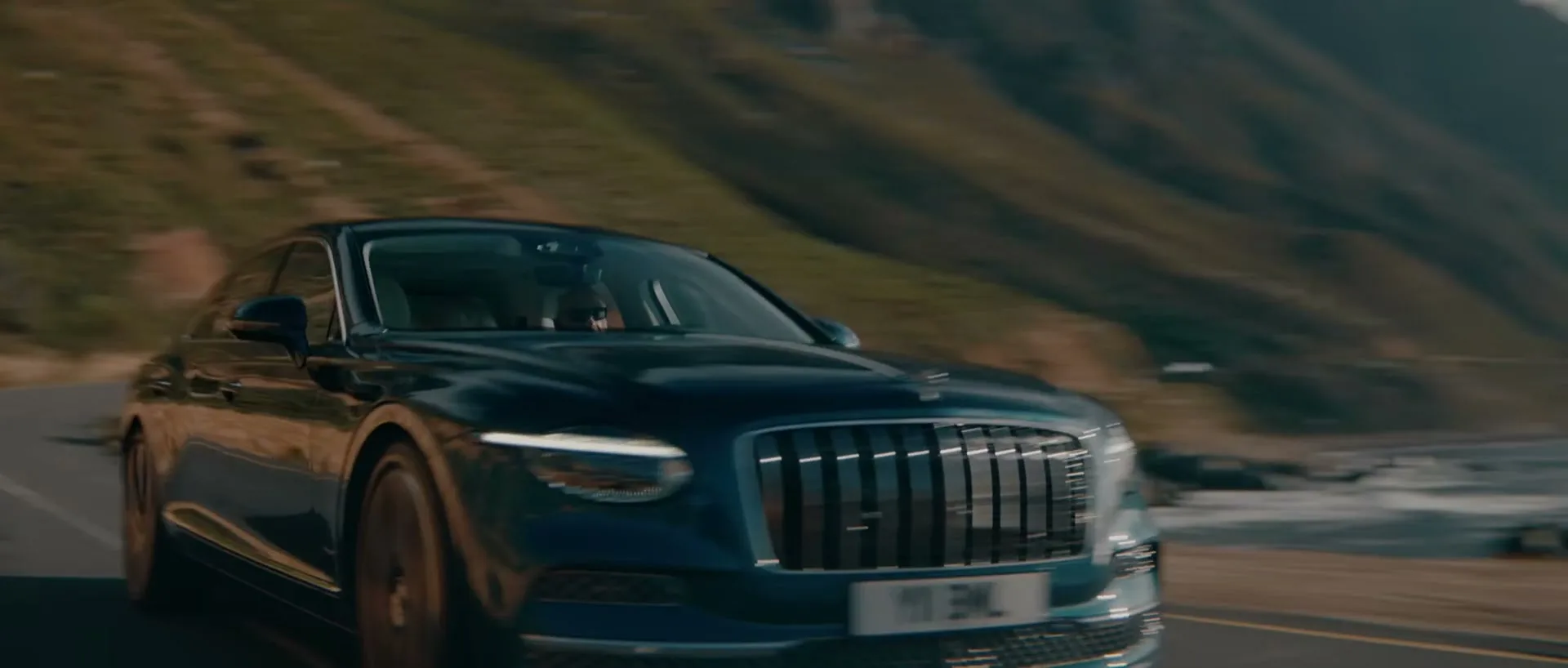 A dark green Bentley car drives along a winding coastal road with mountains and the ocean in the background on a sunny day.