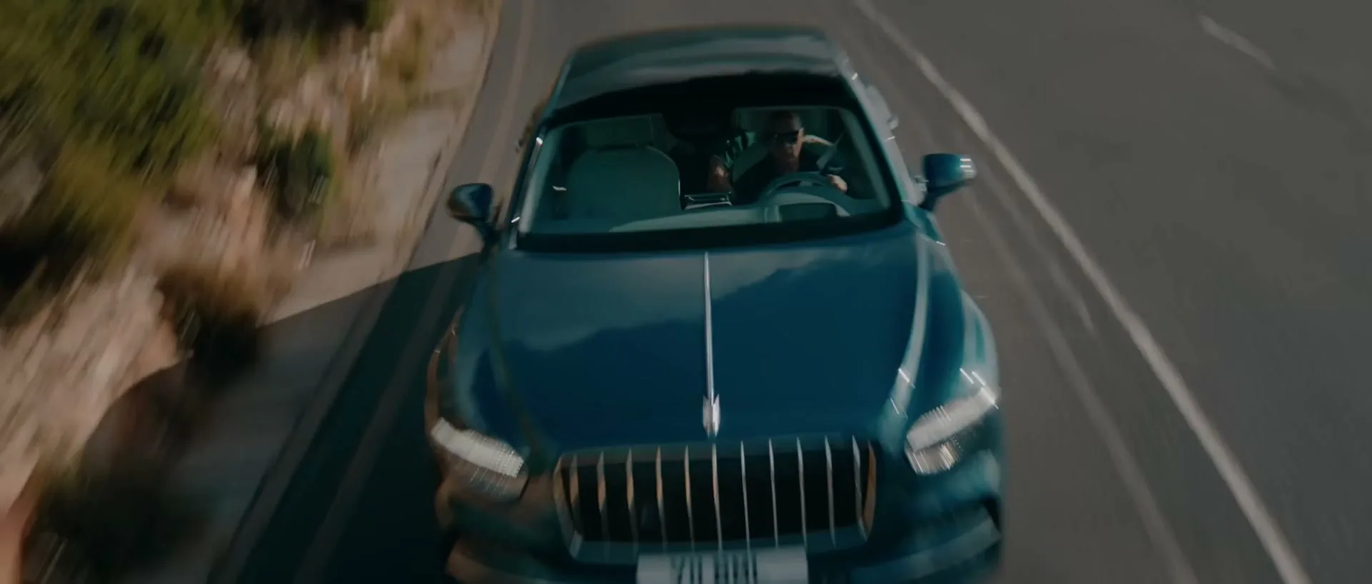 A dark blue Bentley car drives quickly along a winding coastal road during the day, with a male driver visible inside.