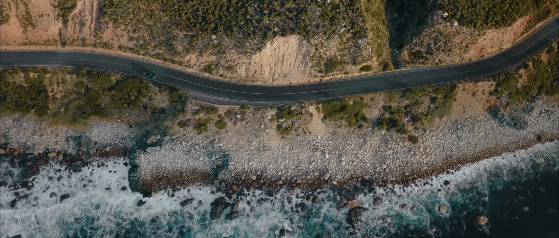 An aerial shot shows a dark car driving along a winding coastal road next to a rocky beach with crashing waves.
