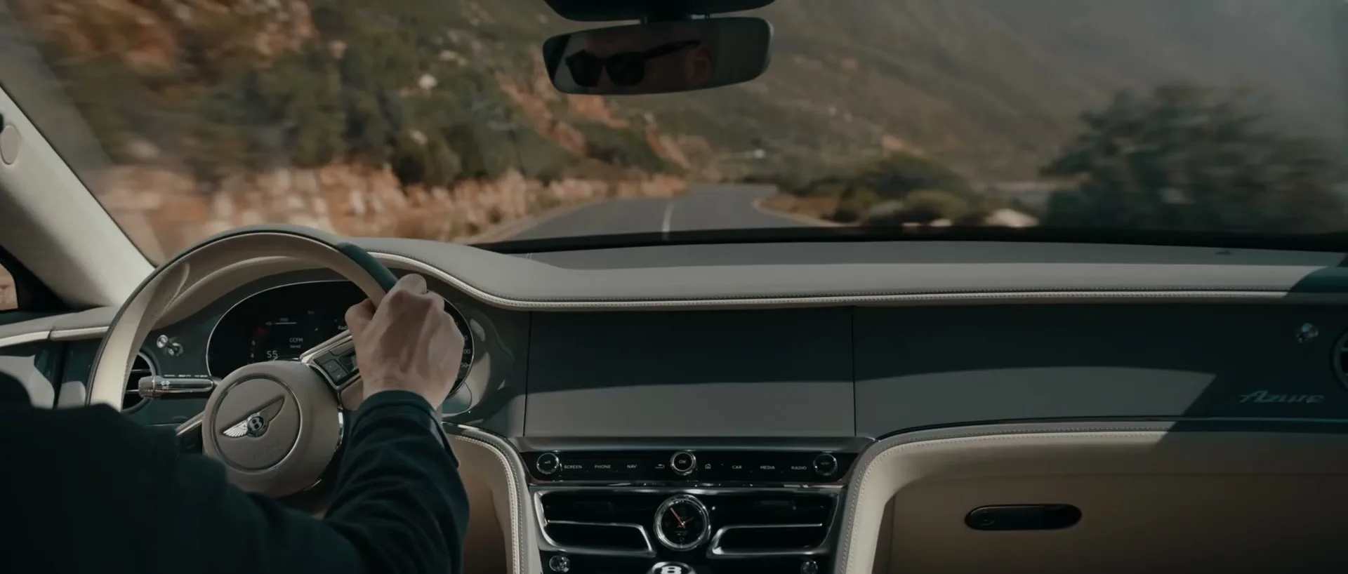 A person's hands are on the steering wheel of a luxury car, driving along a scenic coastal road with mountains and the ocean in the background.