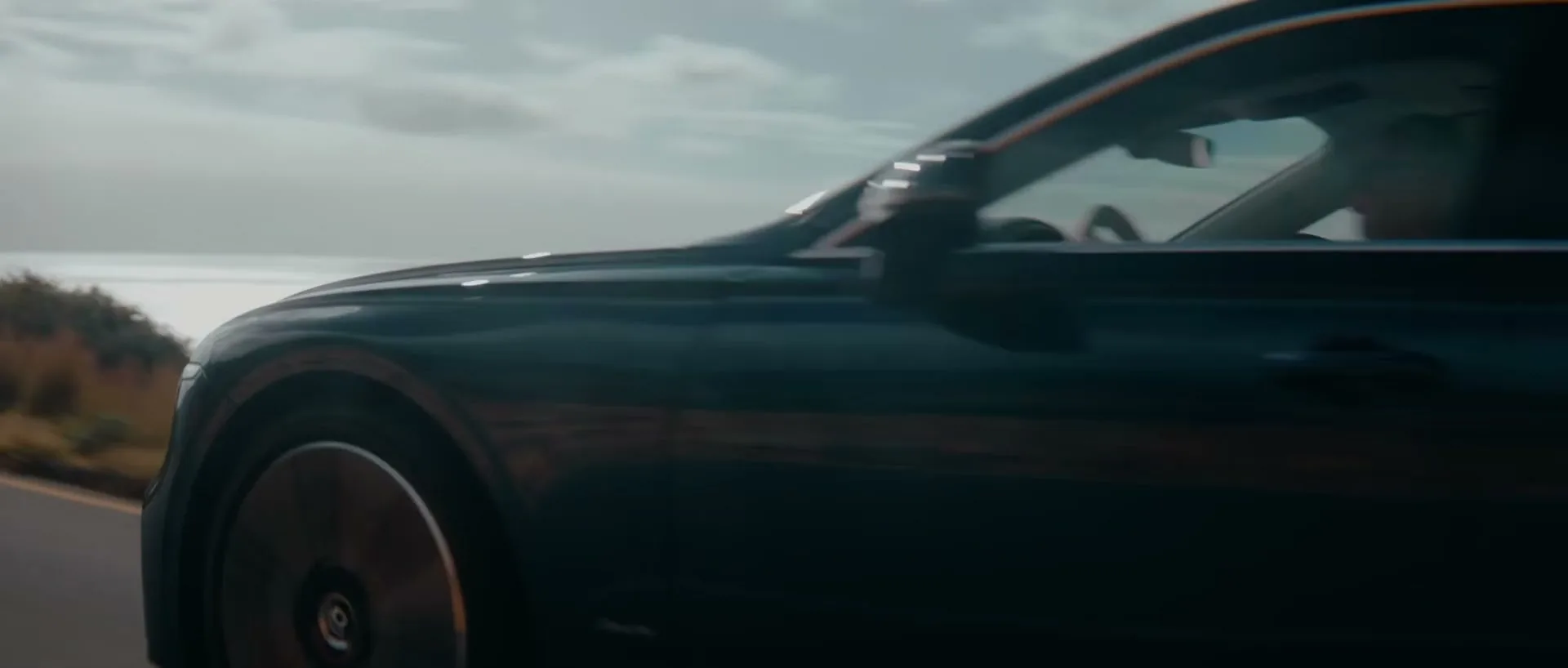 A dark green Bentley car drives along a winding coastal road with mountains and the ocean in the background.