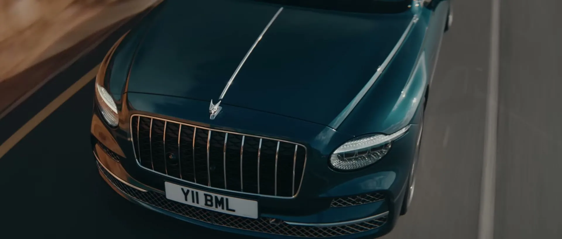 A dark green Bentley car with a chrome grille and headlights drives on a road, viewed from above.