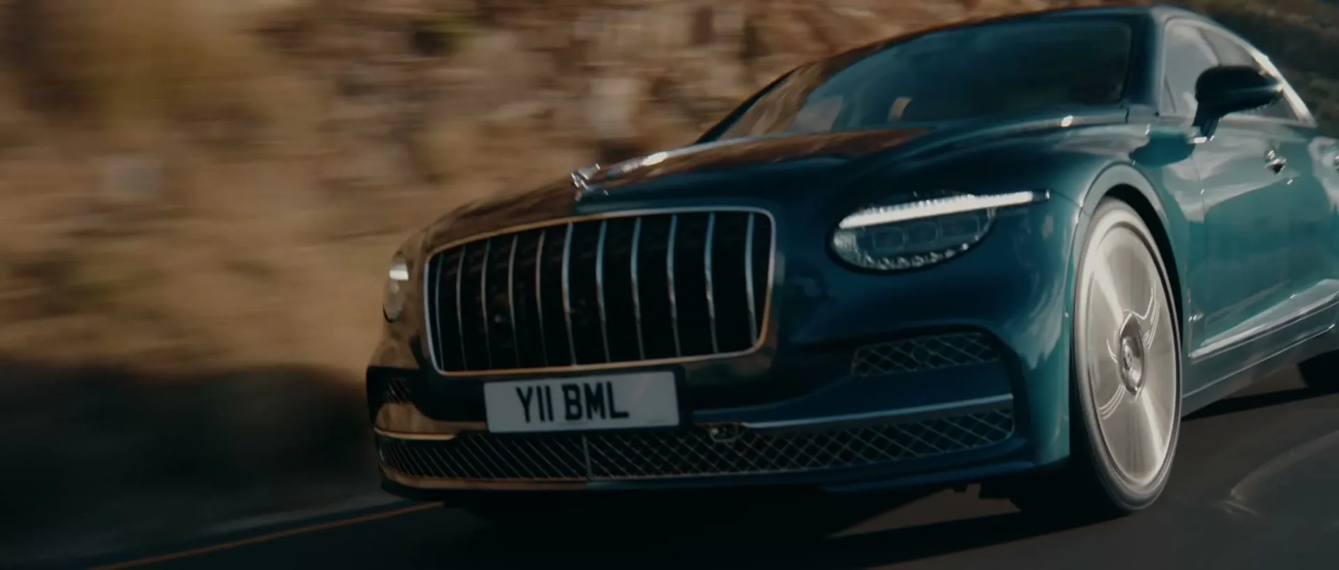 A dark green Bentley car drives quickly along a winding road with mountains in the background during the day.