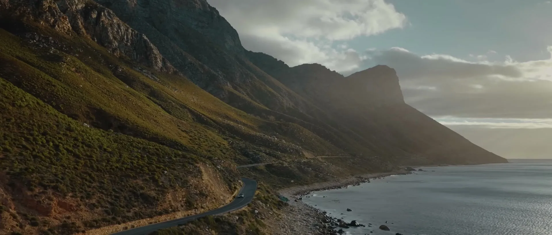 A white Bentley drives along a winding coastal road next to a vast ocean and towering mountains under a cloudy sky.