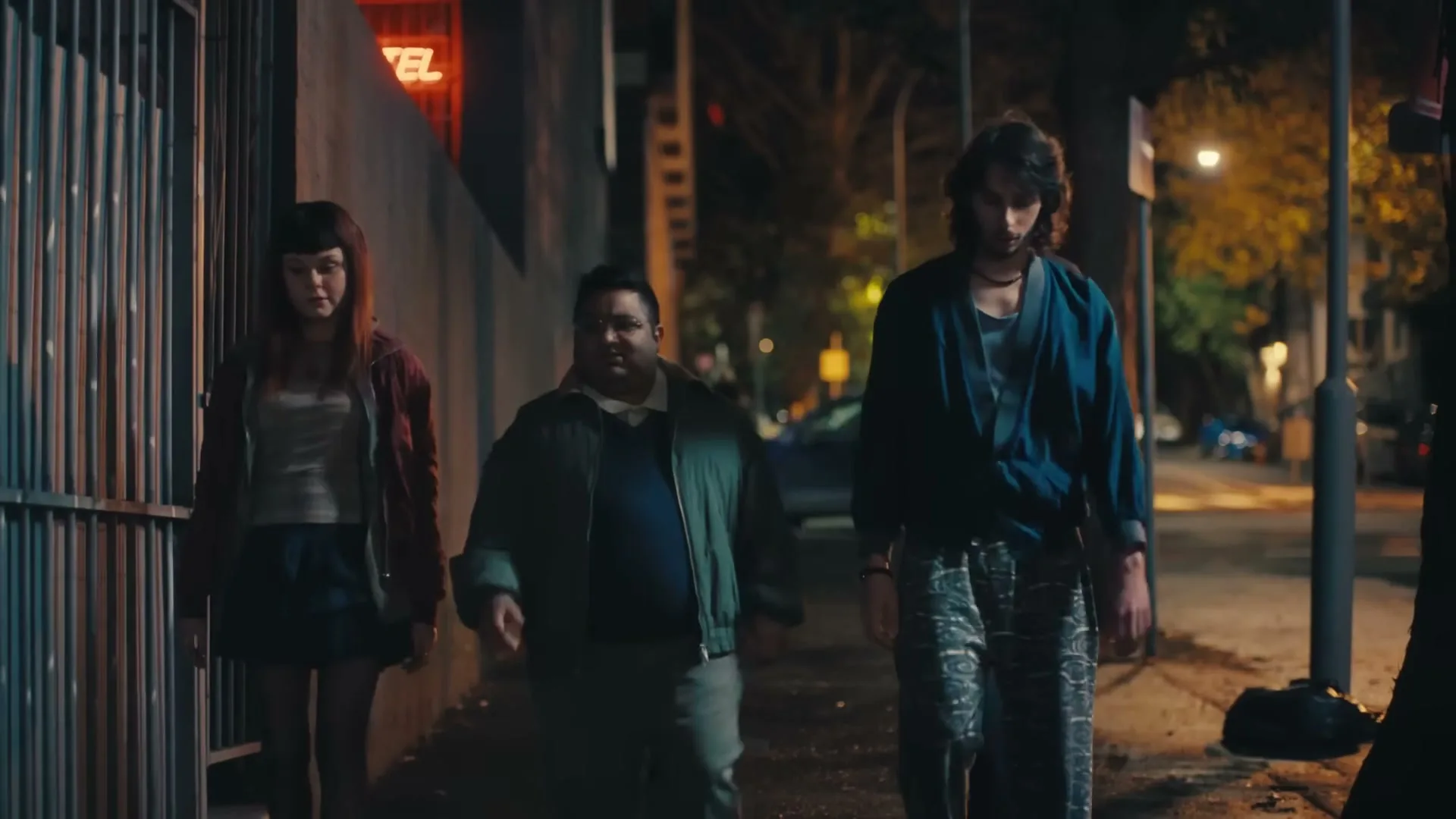 Three friends, a woman, a man, and another man, walk down a city street at night, talking and gesturing.