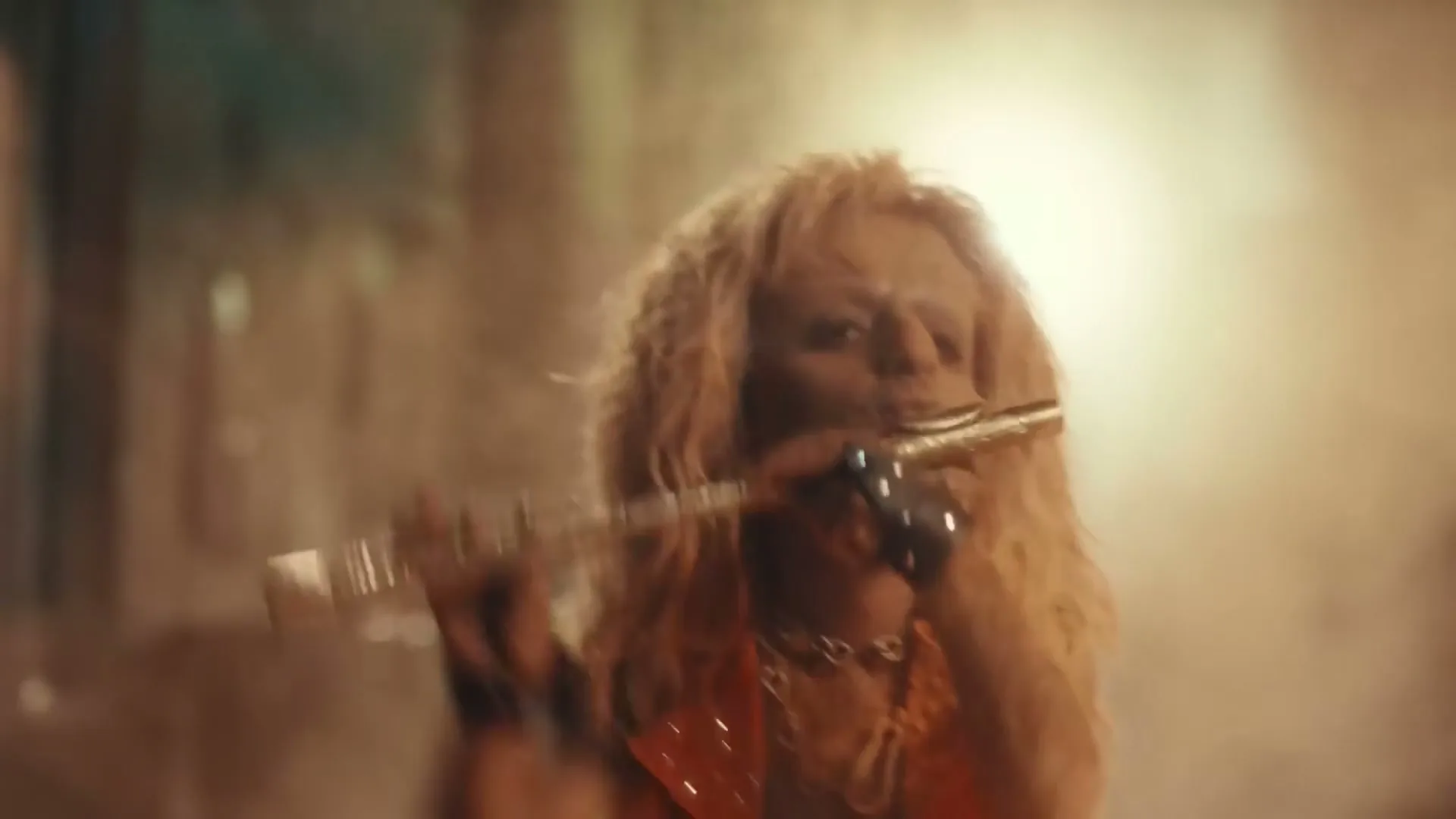 A person with long blonde hair and an orange vest plays a golden flute in a smoky, dimly lit environment.