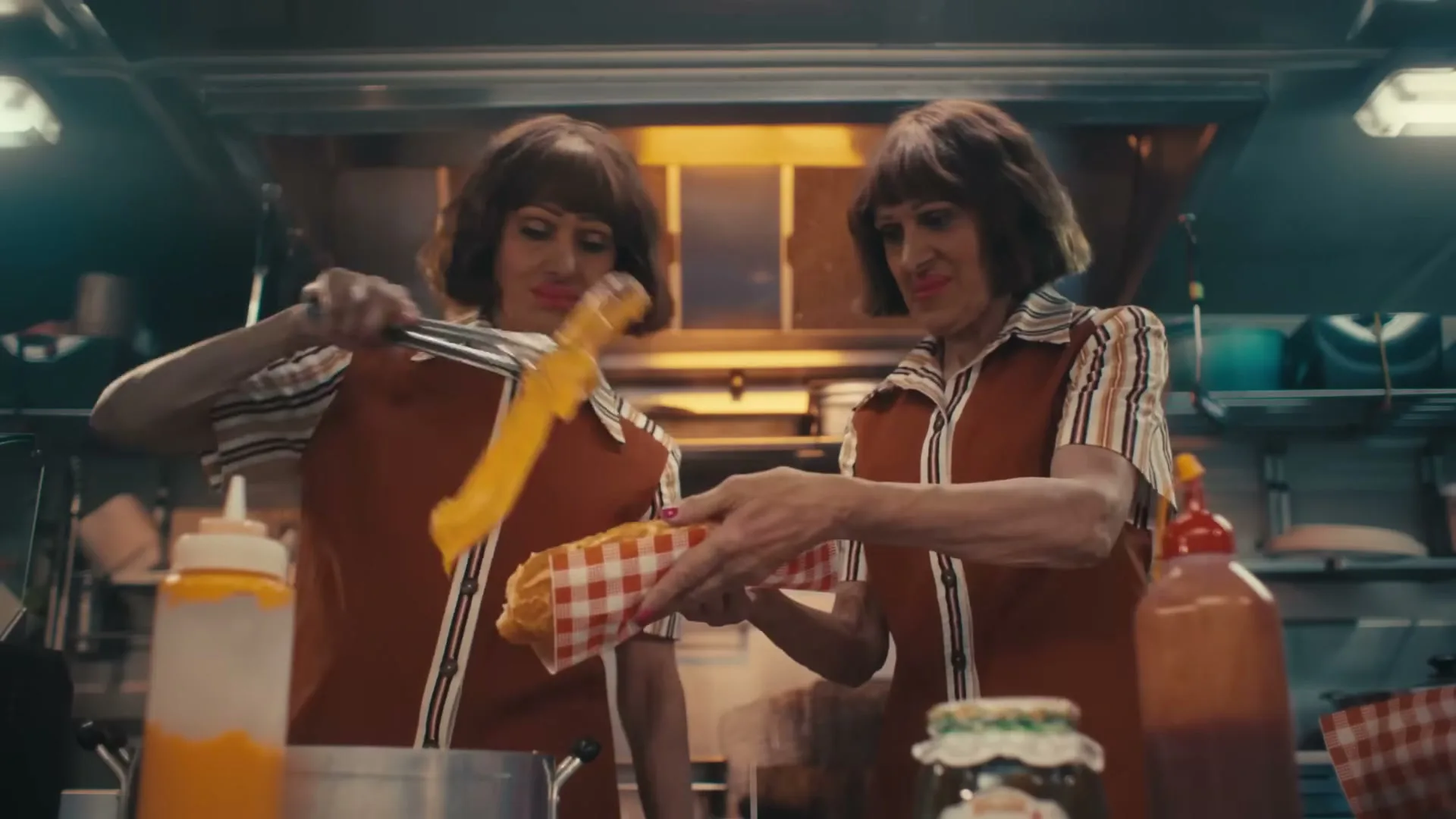 Two identical women in retro uniforms prepare hot dogs in a busy, brightly lit kitchen, one adding cheese and the other holding a wrapped hot dog.