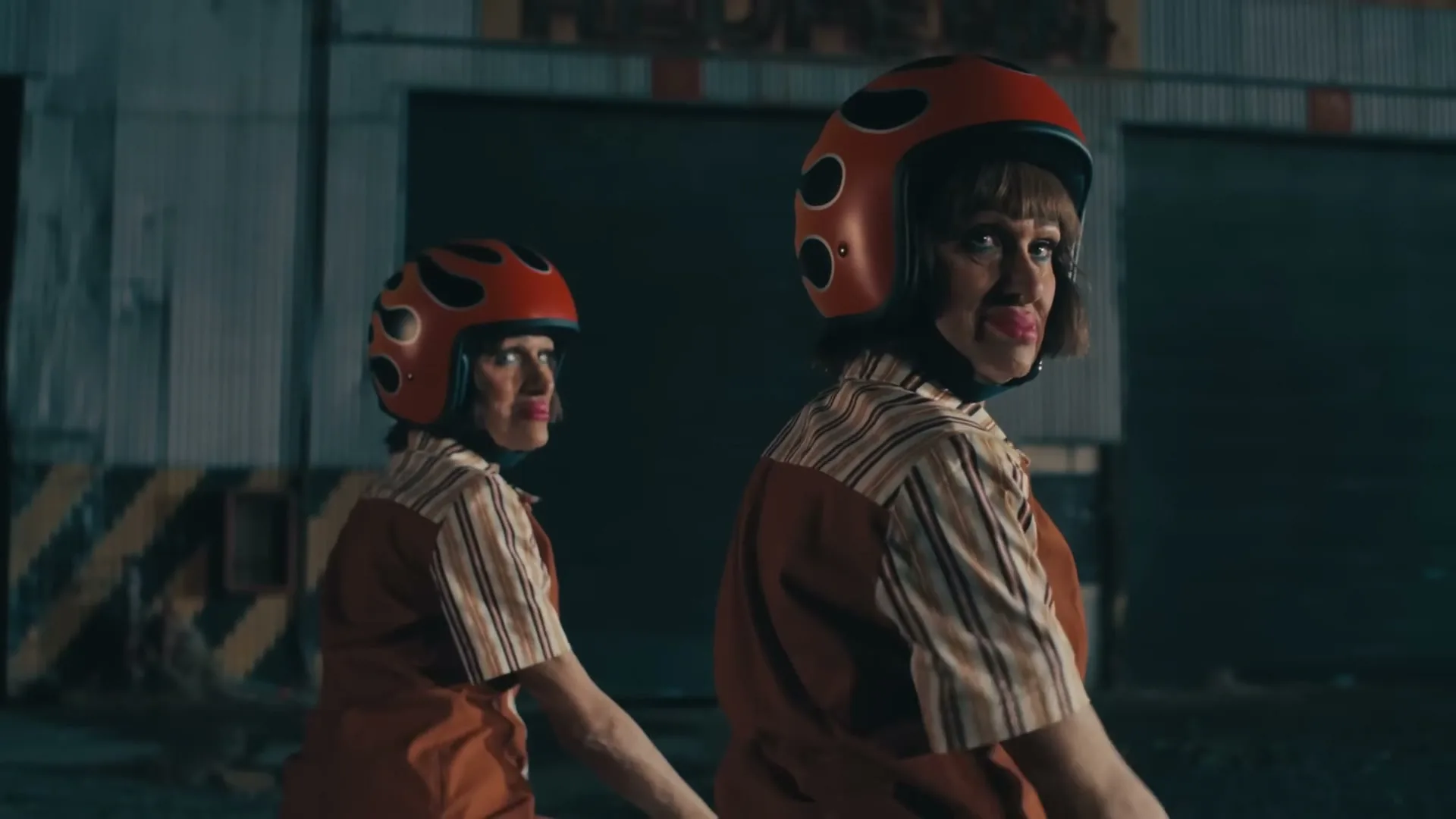 Two women in matching retro outfits and flame-patterned helmets walk away from the camera, then turn and wave in an industrial area at night.