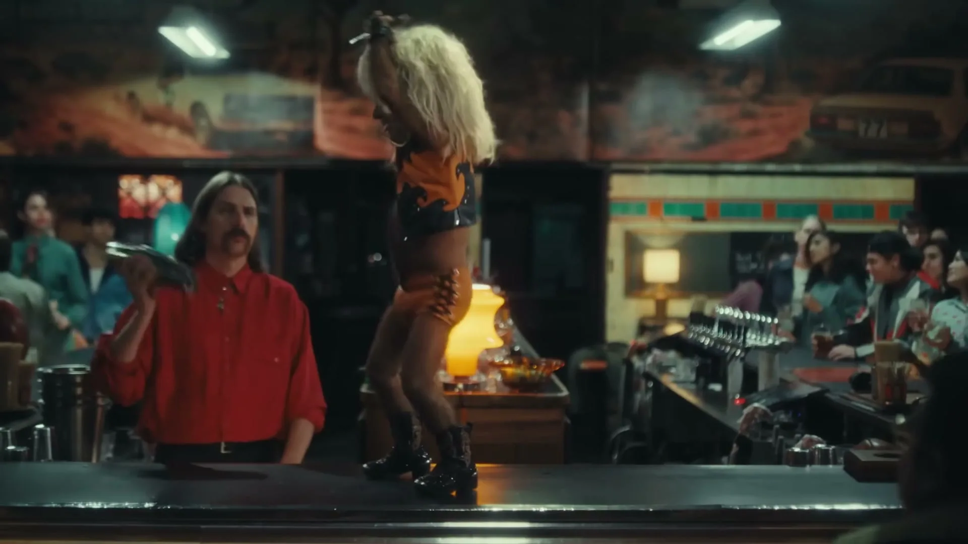 A small person with a blonde wig and orange outfit dances on a bar counter, while a bartender shakes a drink in a lively bar.