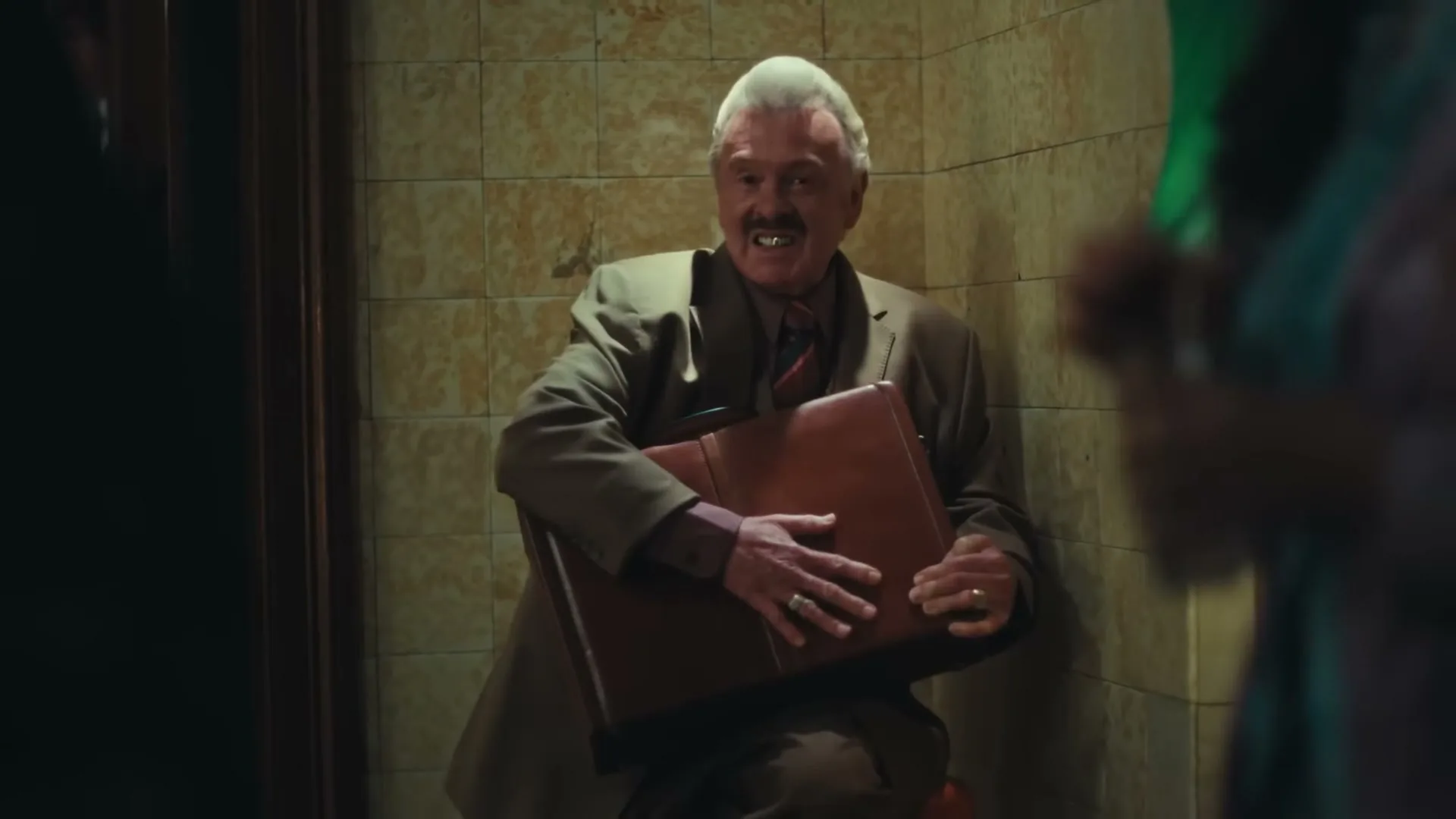 A man with a pompadour and gold teeth, wearing a suit, sits on a stool holding a briefcase and smiling, in a tiled room.