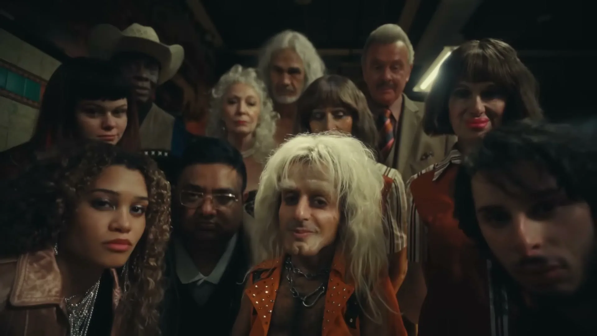 A diverse group of people, some in costume, look down at the camera with varied expressions in a dimly lit interior.