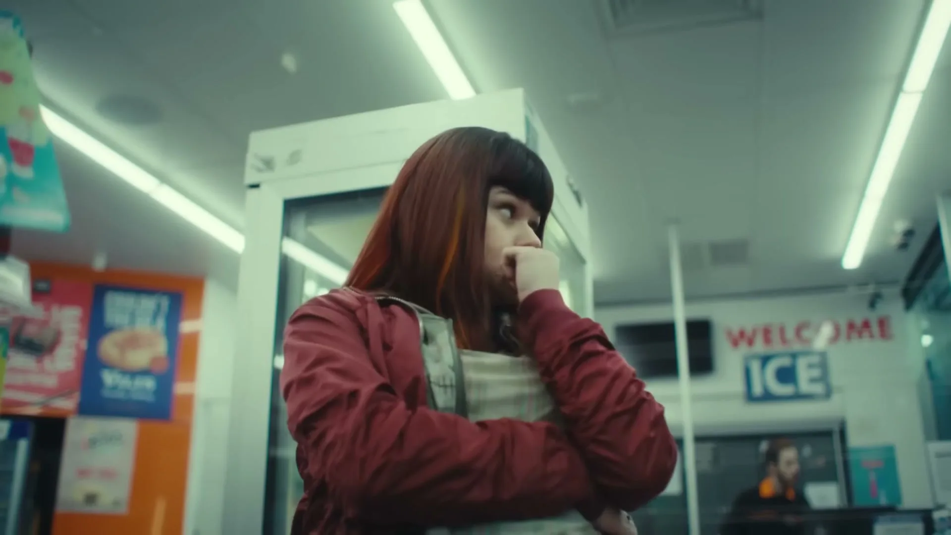 A young woman with red hair and a red jacket looks around nervously in a convenience store.