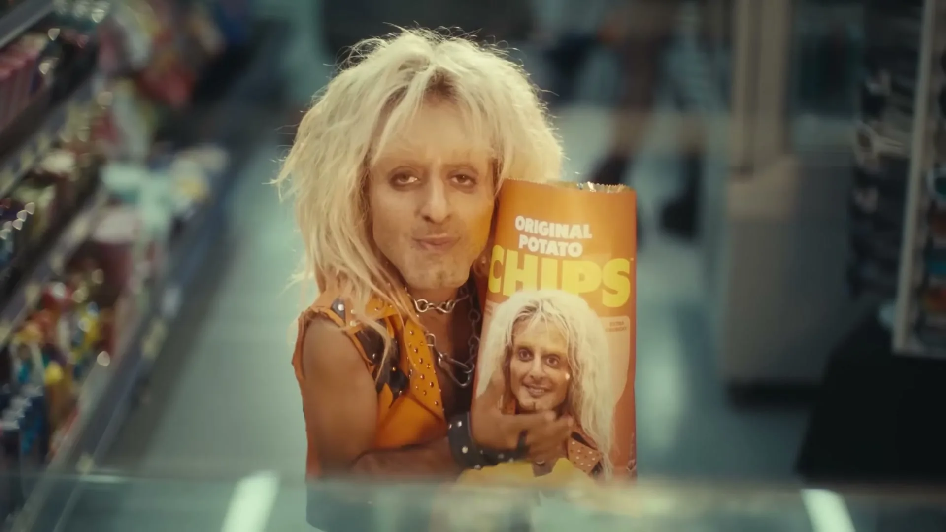 A man with a small head and long blonde hair, wearing a studded vest, holds a bag of potato chips with his face on it, in a grocery store aisle.