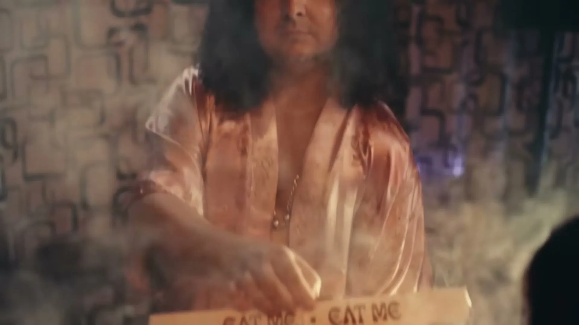 A man with long dark hair and a pink robe holds a pizza box with a steaming pizza and a fried egg on top, in a dimly lit room.
