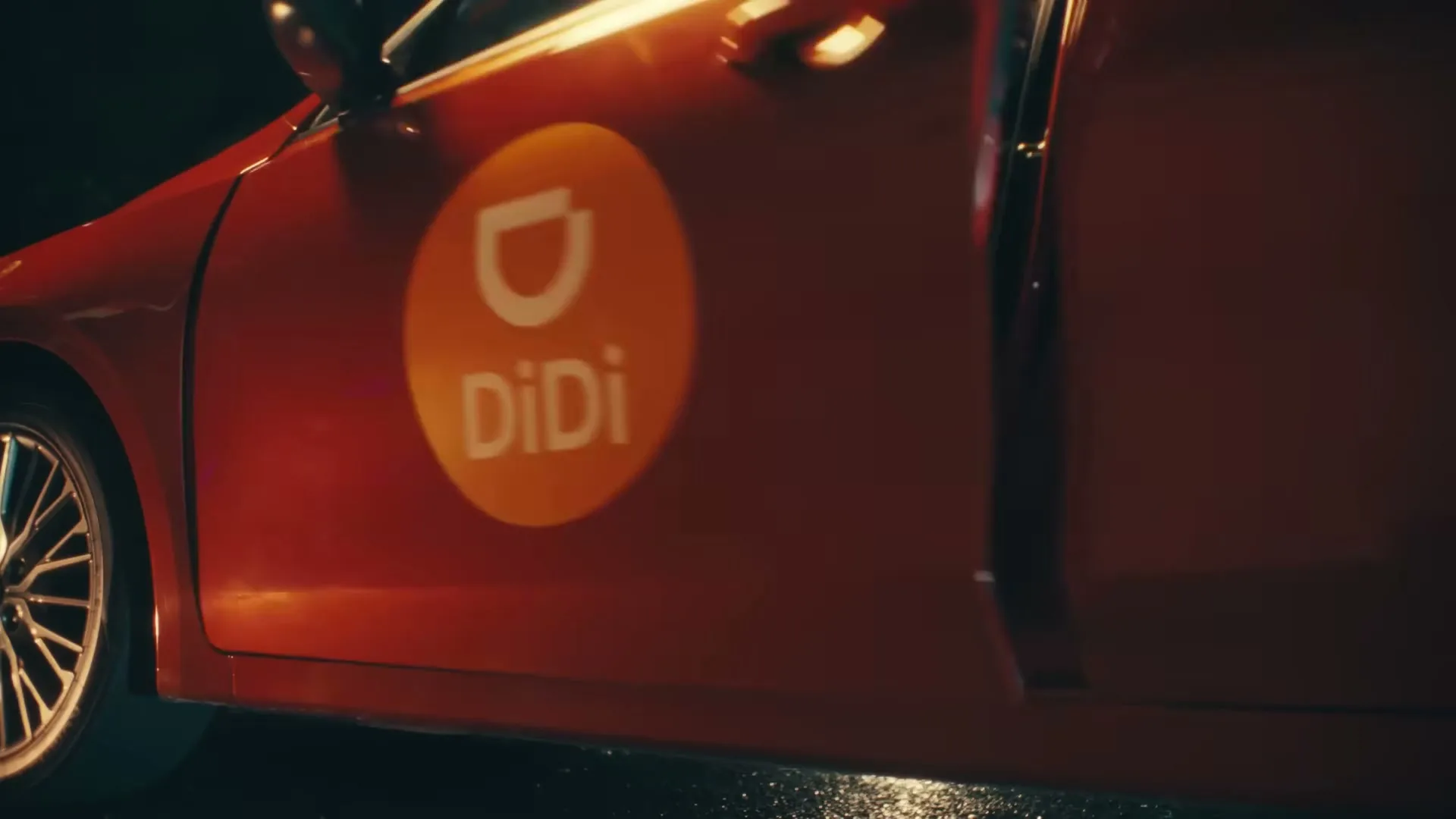 A person in camouflage pants and a blue jacket closes the door of a red car with a DiDi logo on the side at night.